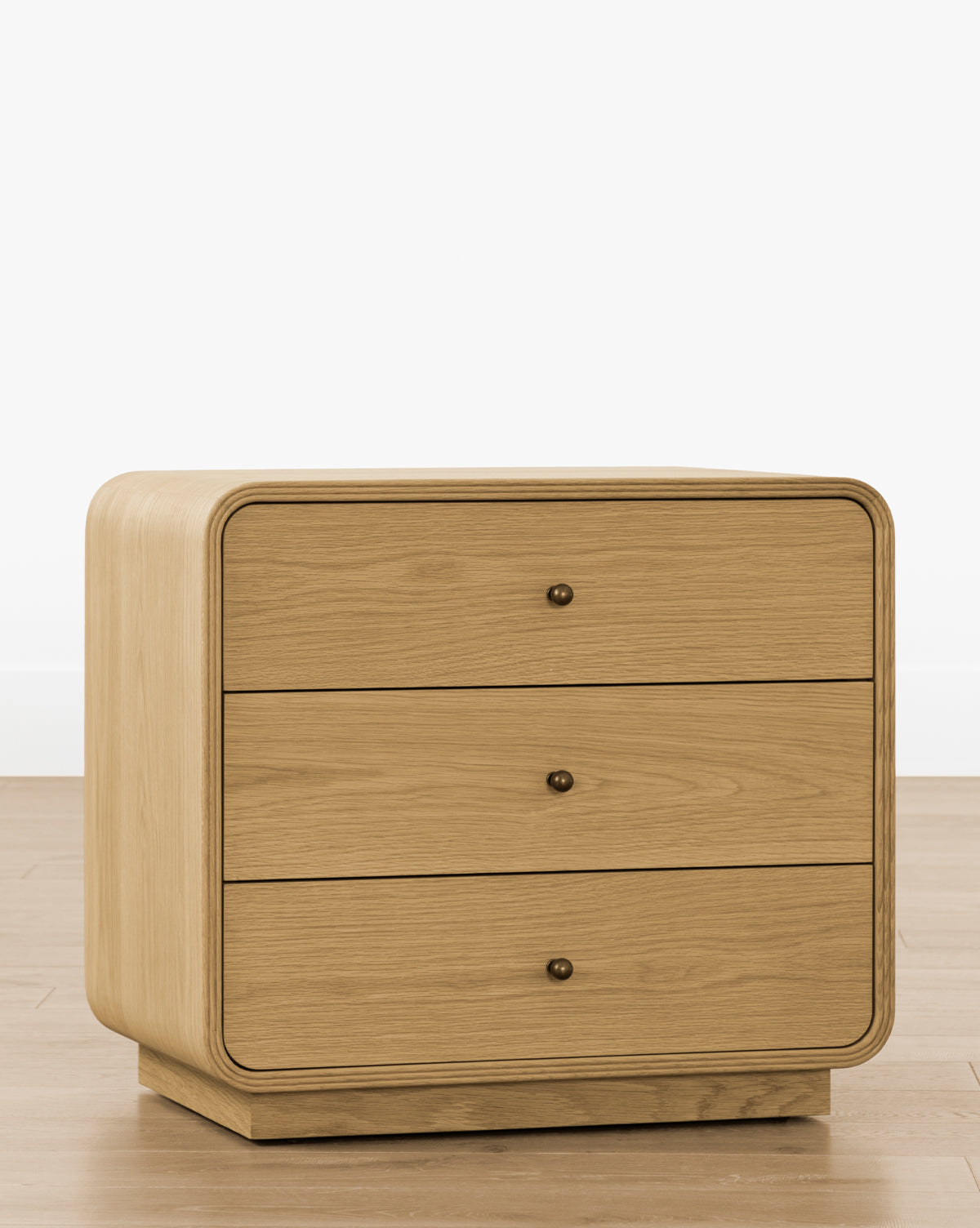 The Wilkins Oak Nightstand, featuring rounded edges and three drawers with round metal knobs, sits on a wooden floor against a plain white background—ideal for enhancing your bedroom decor, by McGee & Co.