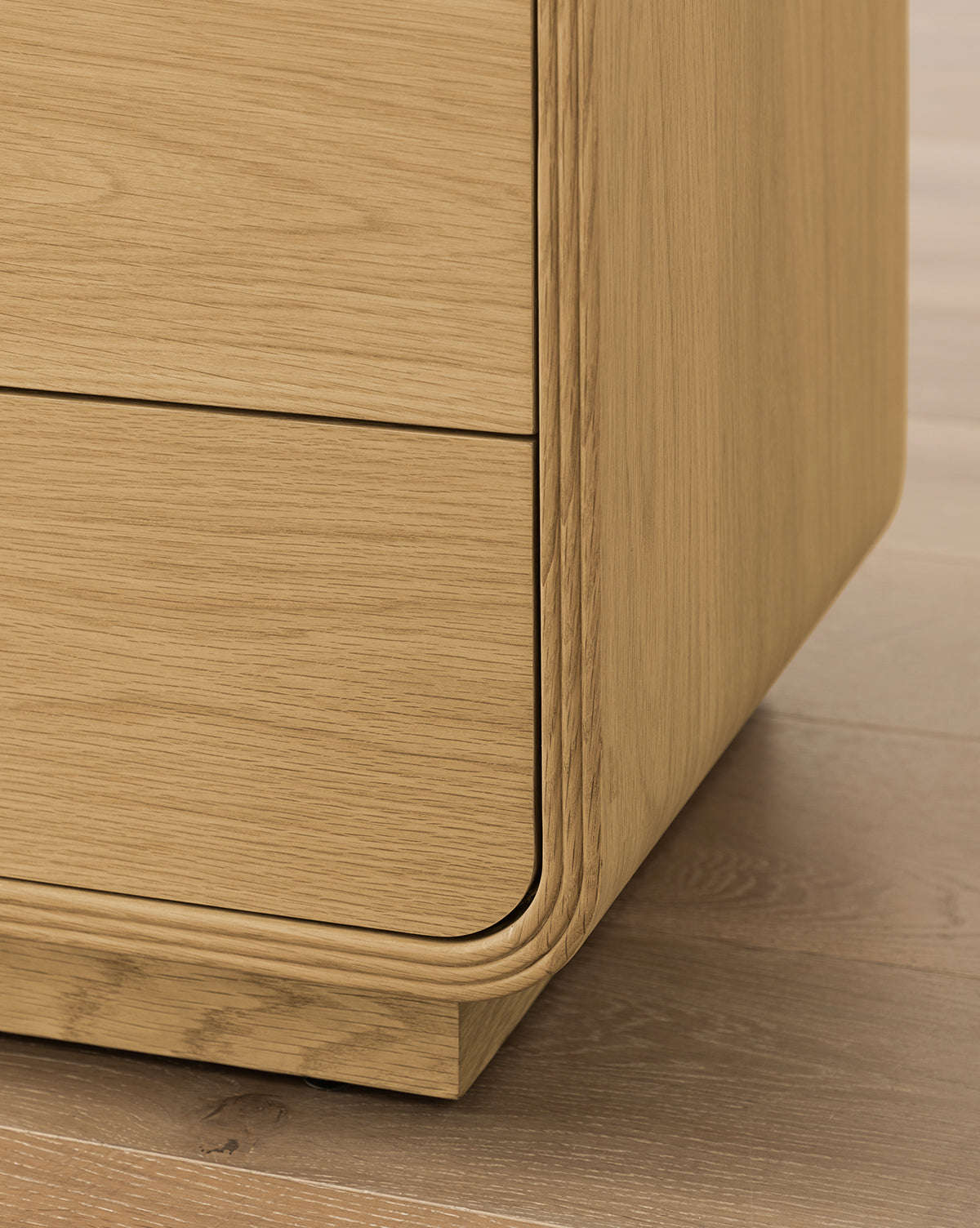 A close-up of the Wilkins Oak Nightstand shows its smooth finish, two drawers, and rounded edges on a wooden floor—an elegant choice to enhance any modern bedroom decor, by McGee & Co.