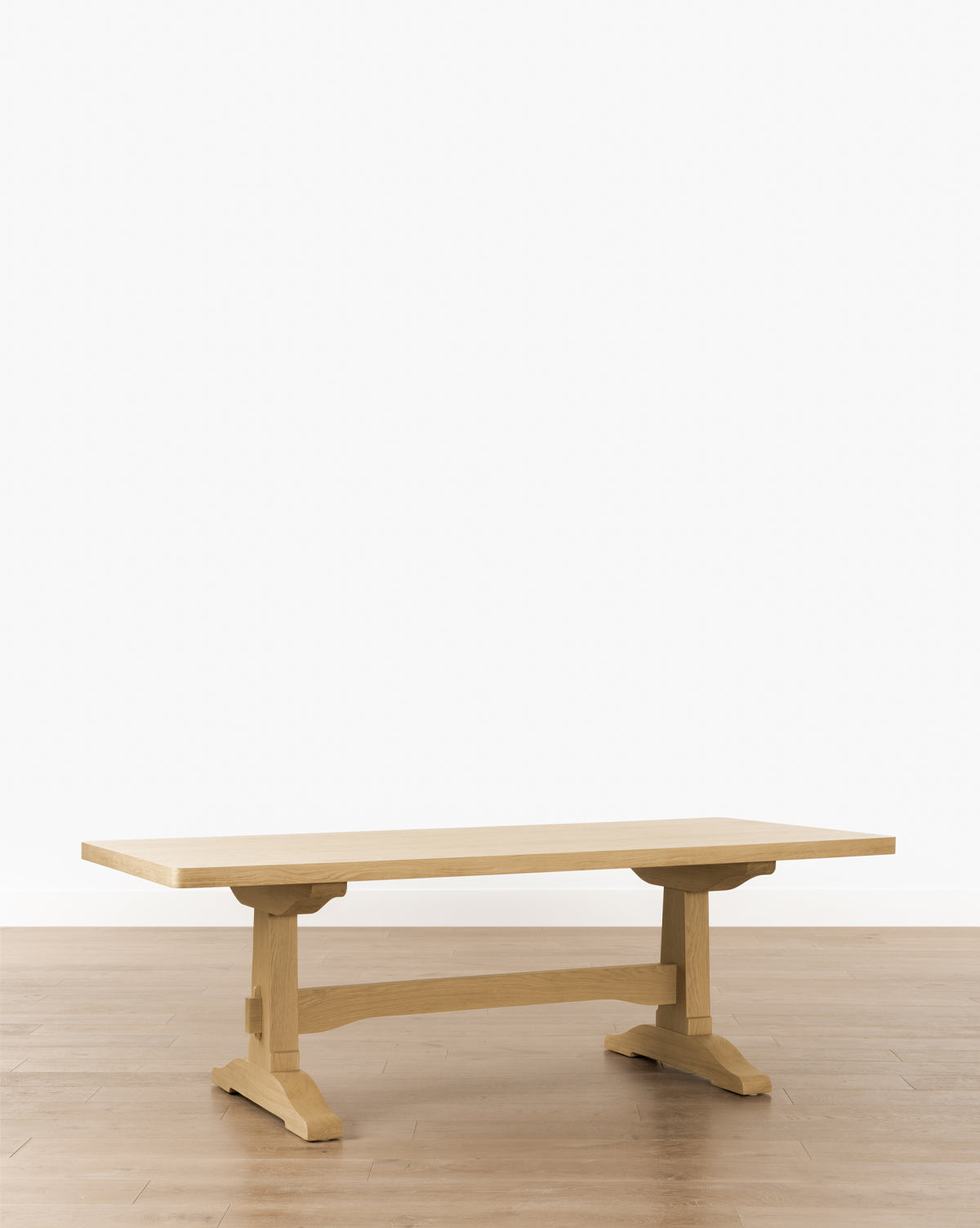 The Wilton Dining Table, featuring a thick rectangular oak top and sturdy trestle legs, is shown on a light brown wooden floor with a plain white background by McGee & Co.