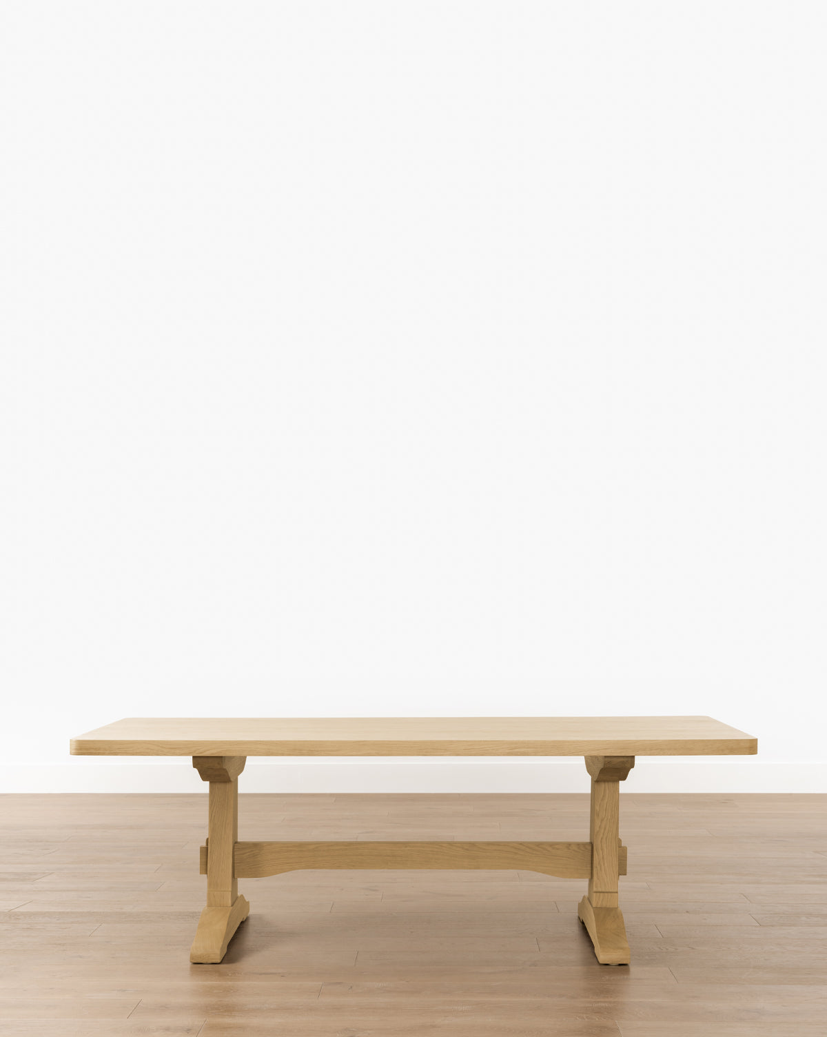 The Wilton Dining Table, featuring a sturdy trestle base and heritage white oak finish, is set on a light brown wooden floor against a plain white wall, by McGee & Co.