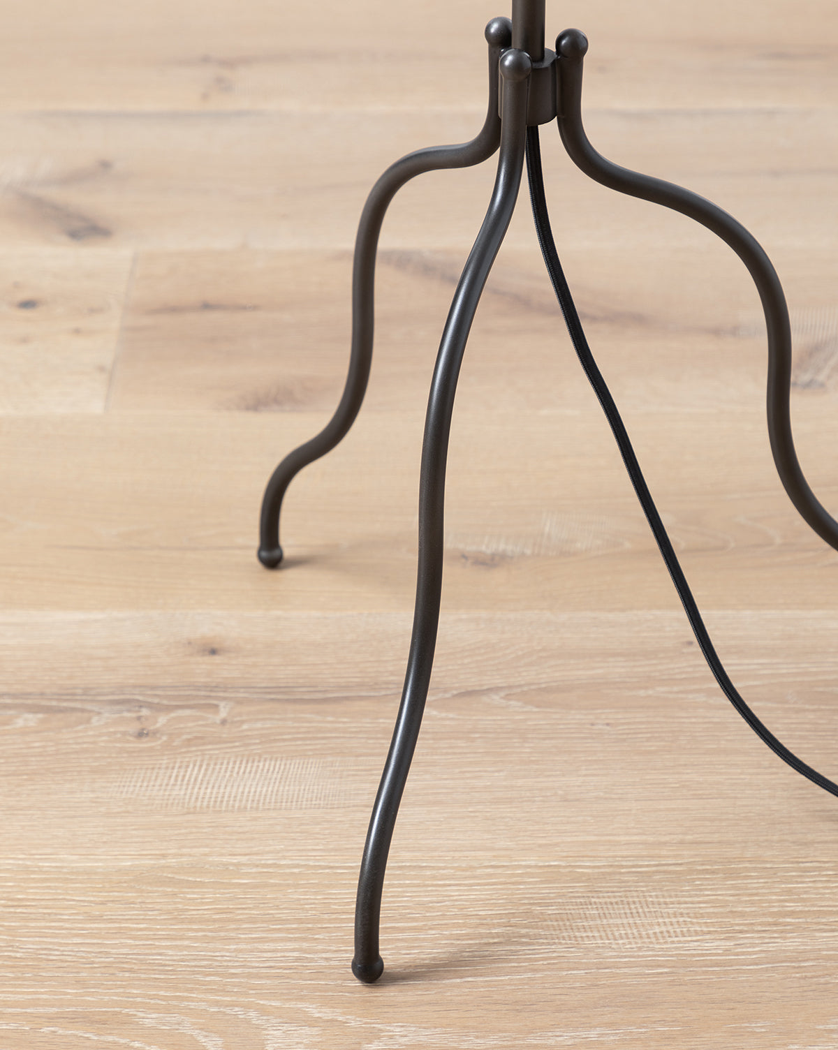 A close-up shows the black metal tripod stand with curved legs of the Windham Floor Lamp on a light wooden floor, its electrical cord wrapped around one leg and a glimpse of the white linen shade above, by McGee & Co.