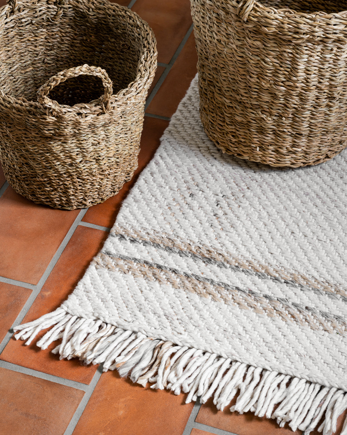 Two woven baskets sit on a terracotta tiled floor beside the Callahan Handwoven Indoor/Outdoor Rug, its neutral colors and fringe adding cozy texture to the space, by McGee & Co.