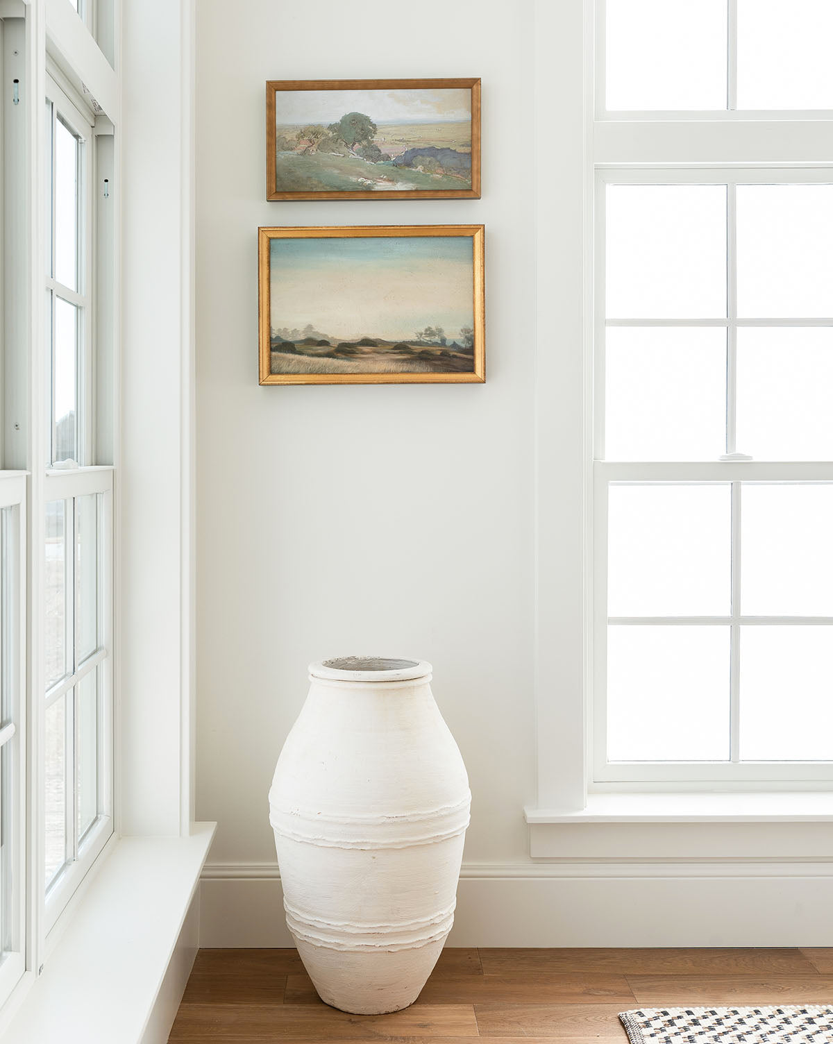 A large white ceramic vase rests on a wooden floor in a bright room. Above it, two framed landscape paintings—including Morning Valley in a gold frame—add character to the space with their scenic beauty.