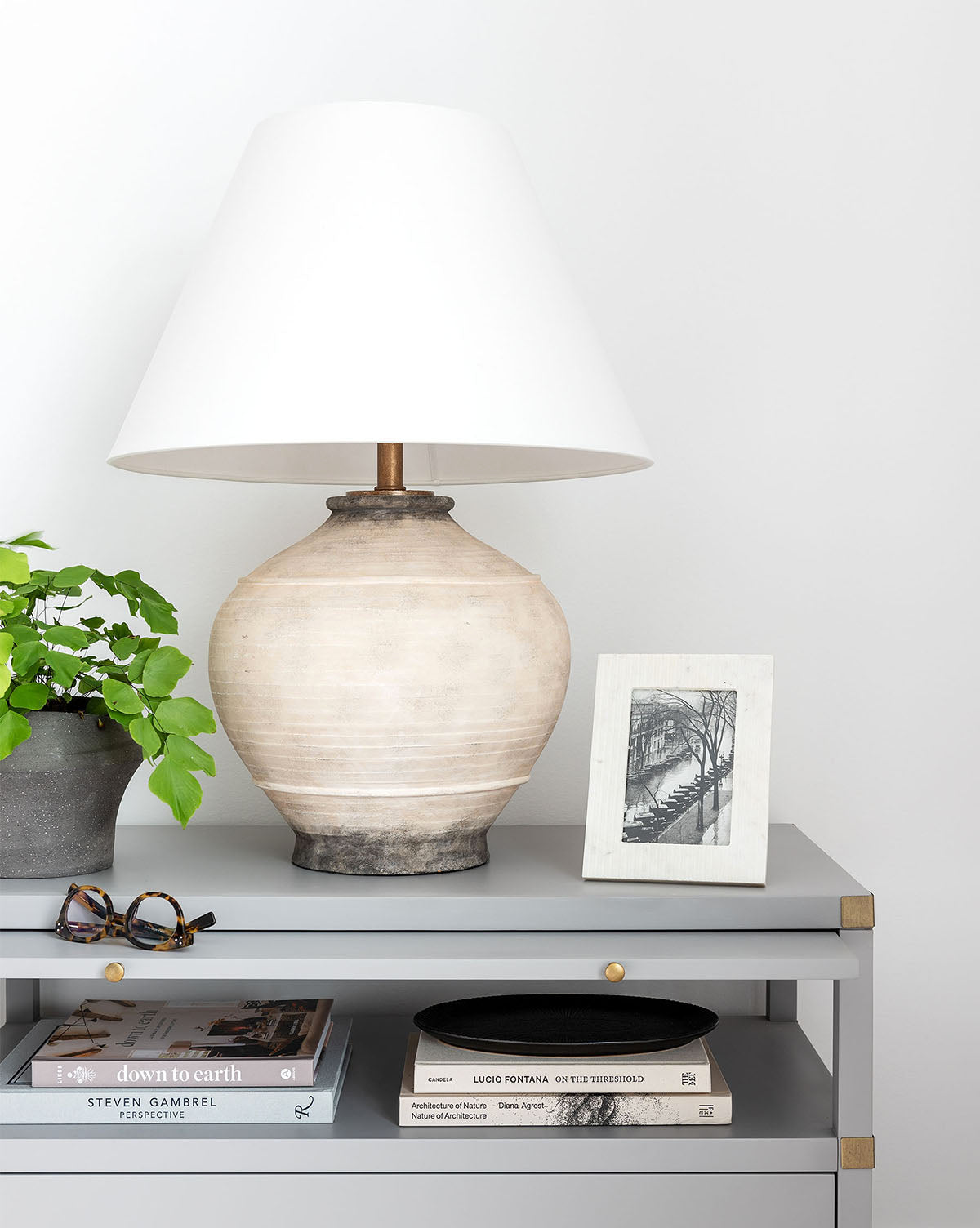 A Malta Table Lamp with a Belgian linen shade sits on a gray console next to a potted plant, black-and-white photo, eyeglasses, and stacked books by McGee & Co.