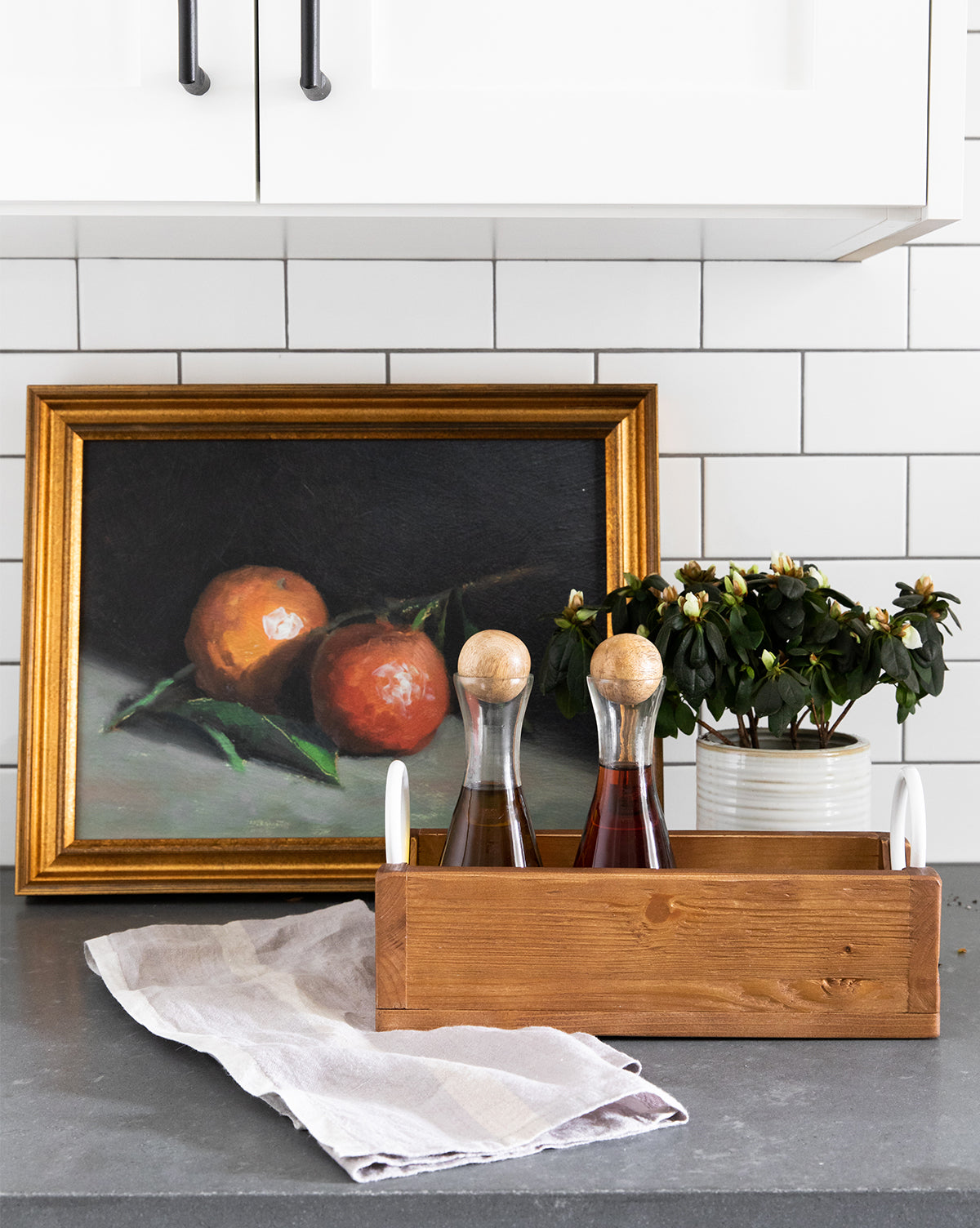 A kitchen countertop displays a wooden tray with glass bottles, a potted plant, a white cloth, and the framed artwork Still Life with Oranges, creating a charming gallery wall look against white subway tile.
