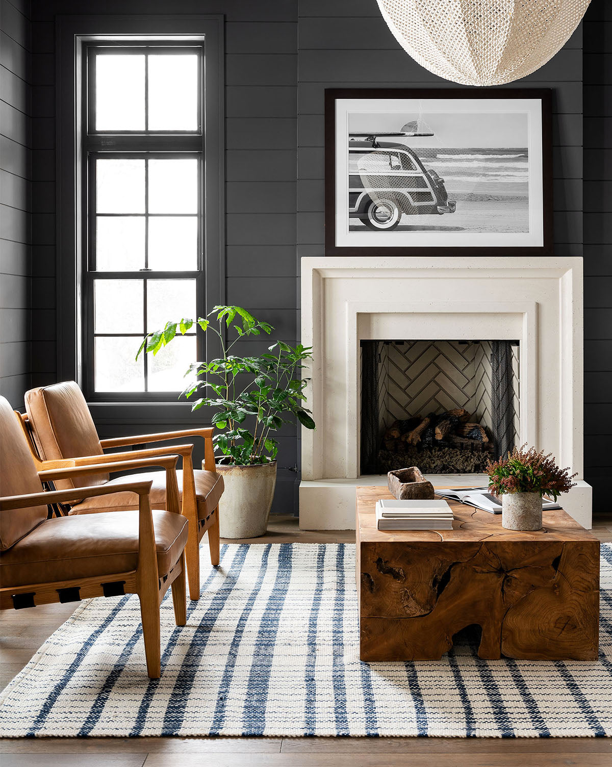 A modern living room with a white fireplace, wooden chairs, tan cushions, coffee table, coastal cool decor, plants, and Gone Surfin black and white wall art above the mantel.