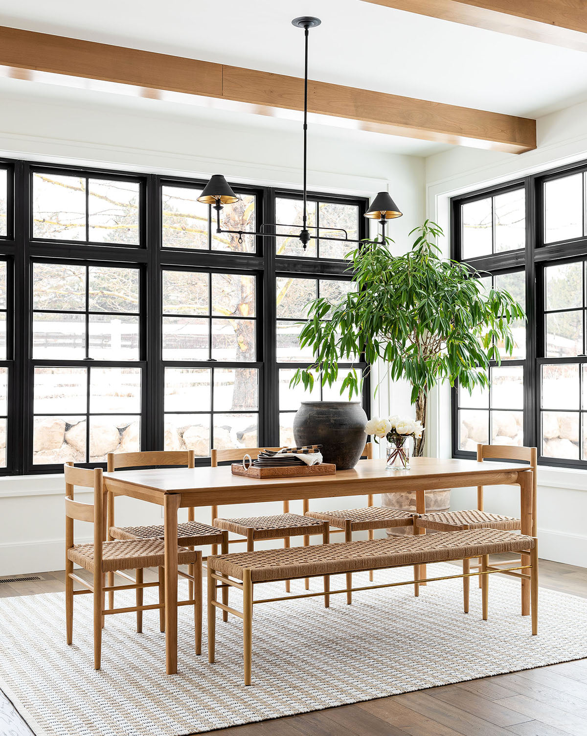 A modern dining room features large black-trim windows, the sturdy Alec Dining Table, woven chairs, a bench, a striped rug, two black pendant lights, and a big potted plant under wooden ceiling beams by McGee & Co.