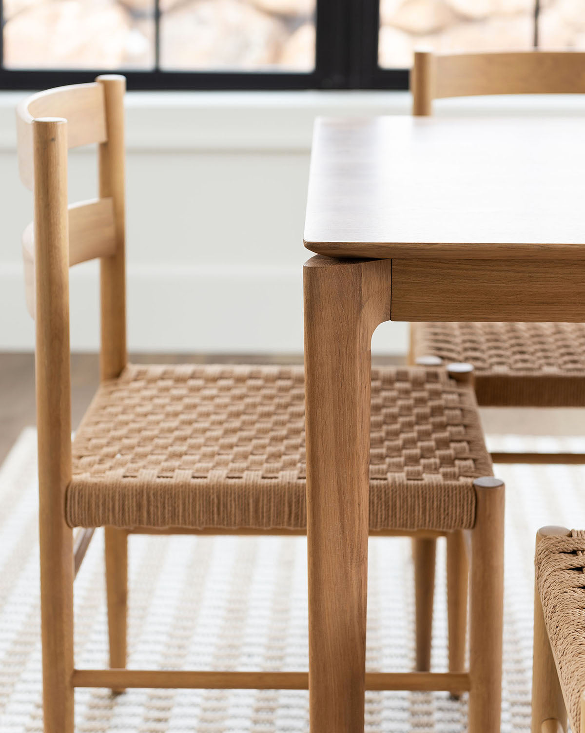 A close-up of the sturdy Alec Dining Table and two matching woven-seat chairs, set on a textured white rug by a black-trimmed window—perfect for any dining area, by McGee & Co.