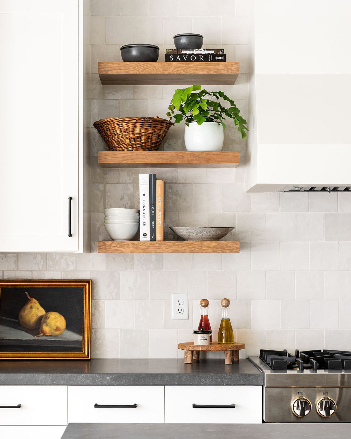 Modern kitchen with floating wooden shelves displaying dishes, books, a wicker basket, and a potted plant. On the counter below are oil and vinegar bottles, a gas stove on the right, and the Still Life with Pears portrait in a frame.