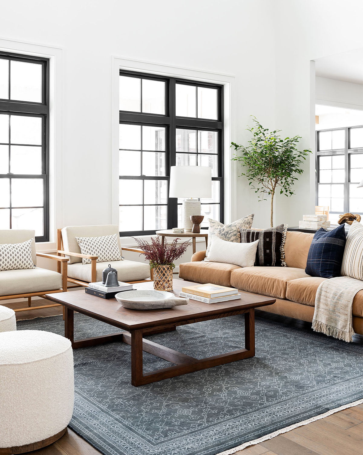 A bright, modern living room with large black-framed windows, a tan sofa, two beige armchairs, the Herrin Coffee Table, a gray patterned rug, decorative pillows, and a potted plant in the corner, by McGee & Co.