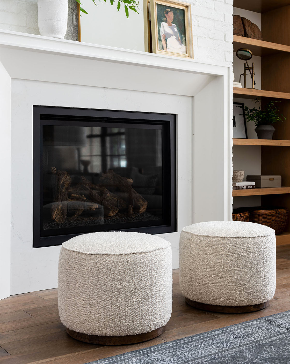 Two round Dawson Ottomans with textured fabric cushions sit before a modern white fireplace with a black frame. Wooden shelves and decorative accents, including framed art, enhance the inviting background by McGee & Co.