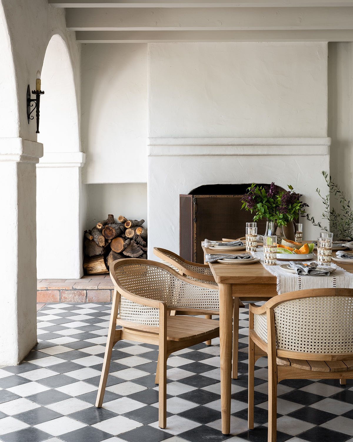 A dining area features Elowyn Outdoor Dining Chairs around a wooden table, set for a meal on black and white checkered tiles, with a fireplace, stacked firewood, and archways in a bright, white-walled room by McGee & Co.