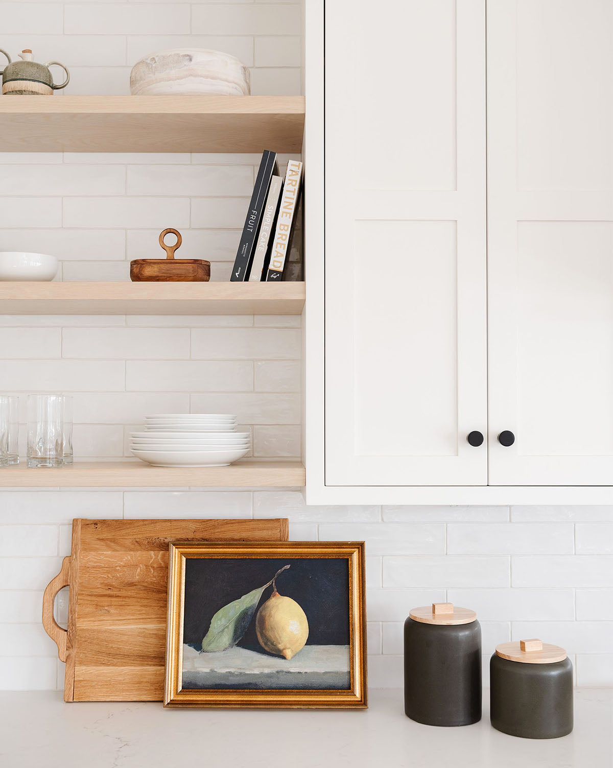 A kitchen counter features two dark jars, a wooden cutting board, and the Still Life with Lemon framed painting. Open shelves above display dishes, glasses, books, and decor beside white cabinets.