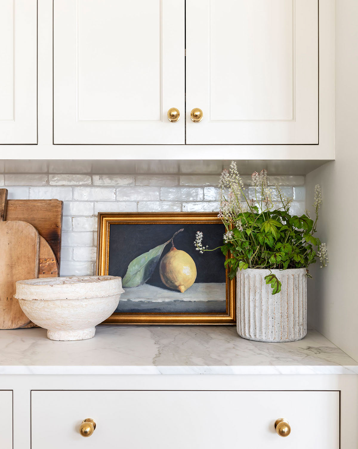 A kitchen countertop features a white bowl, the “Still Life with Lemon” framed art, a potted green plant, and wooden cutting boards set against a white tile backsplash and white cabinets with gold knobs.