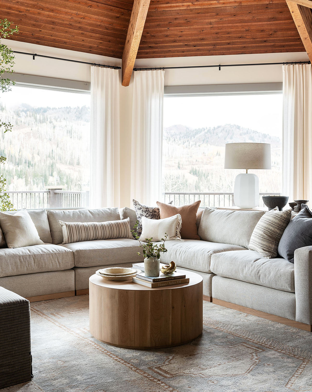 A bright living room with large windows showing a mountain view, the Vernon Modular L Sectional 117.5 with assorted throw pillows, a round wooden coffee table with decor, and a neutral rug under a wood-beamed ceiling, by McGee & Co.