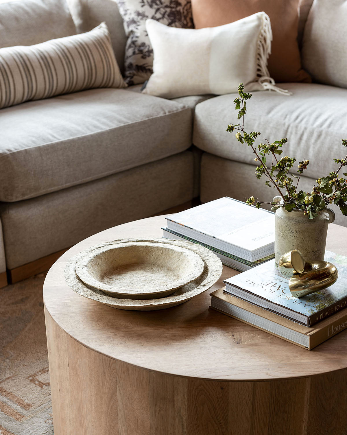 A round wooden coffee table with stacked books, two textured shallow bowls, and the Dual Handled Vase with green branches sits in front of a beige sectional sofa accented by throw pillows.