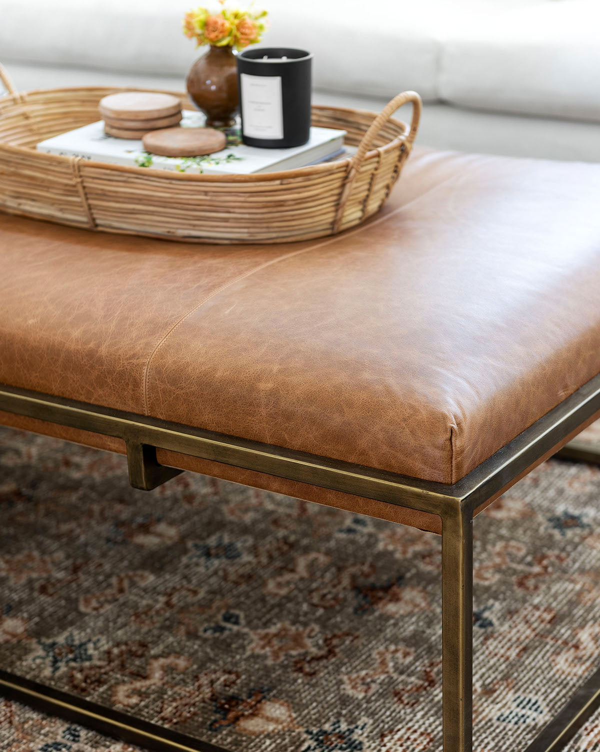The Harlow Leather Coffee Table features an aniline-dyed rectangular leather top with a modern brass-finished frame. A woven tray holds a black candle, coasters, and yellow flowers. The ottoman rests on a patterned rug, by McGee & Co.
