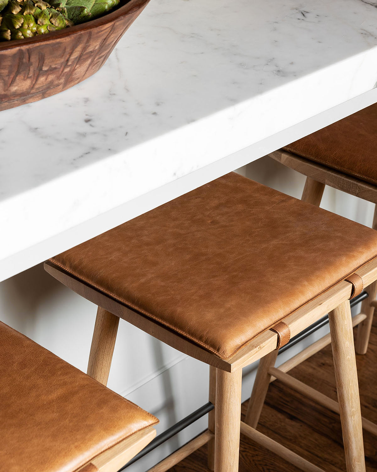 A close-up of tan leather-cushioned Beau Counter Stools under a white marble countertop, with a wooden bowl of artichokes partially visible on the counter, by McGee & Co.