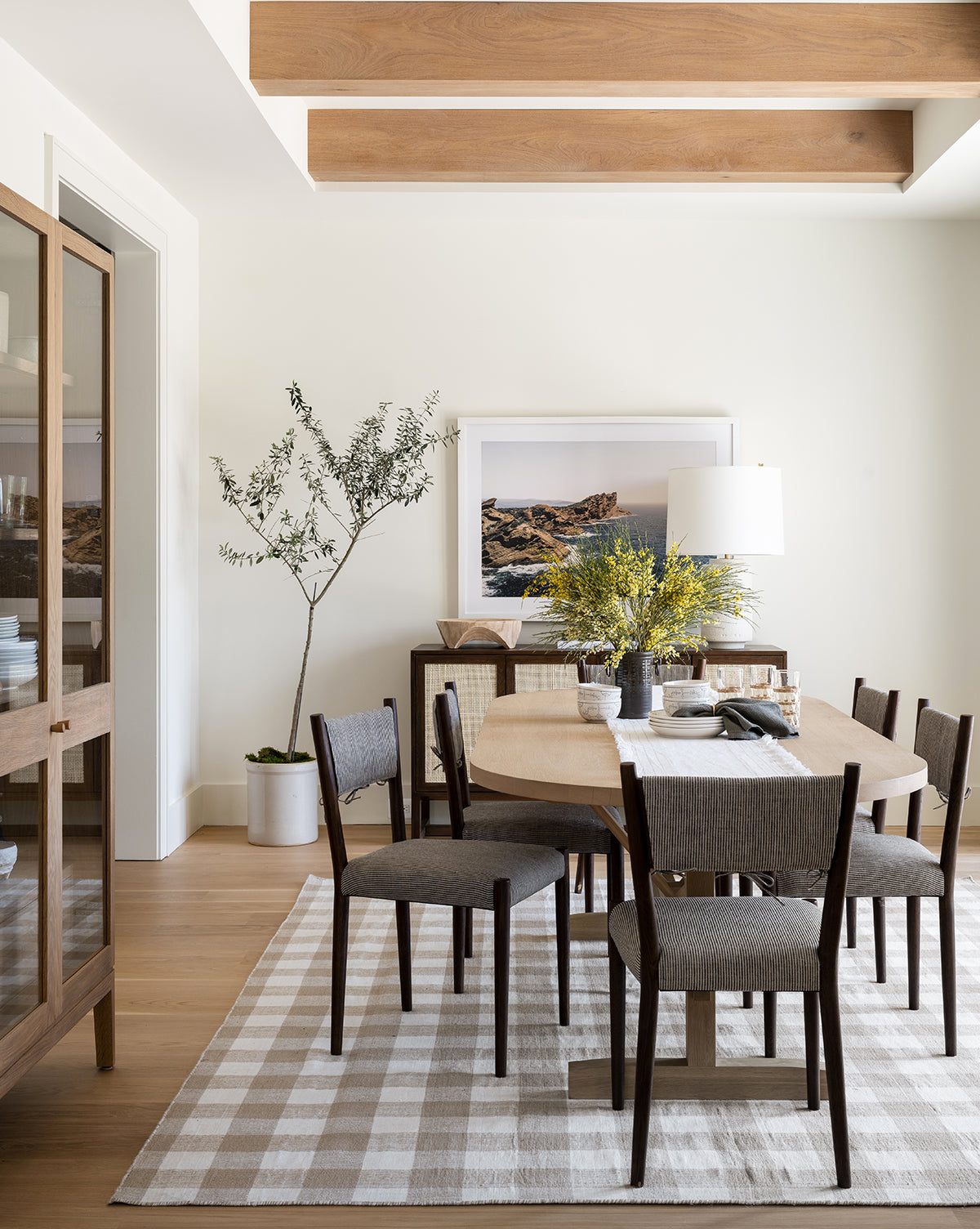 A modern dining room with a Giselle Oval Dining Table, six gray chairs, a patterned rug, potted plant, and sideboard topped with a lamp, yellow flowers, and framed art. Exposed wood ceiling beams complete the cozy decor, by McGee & Co.