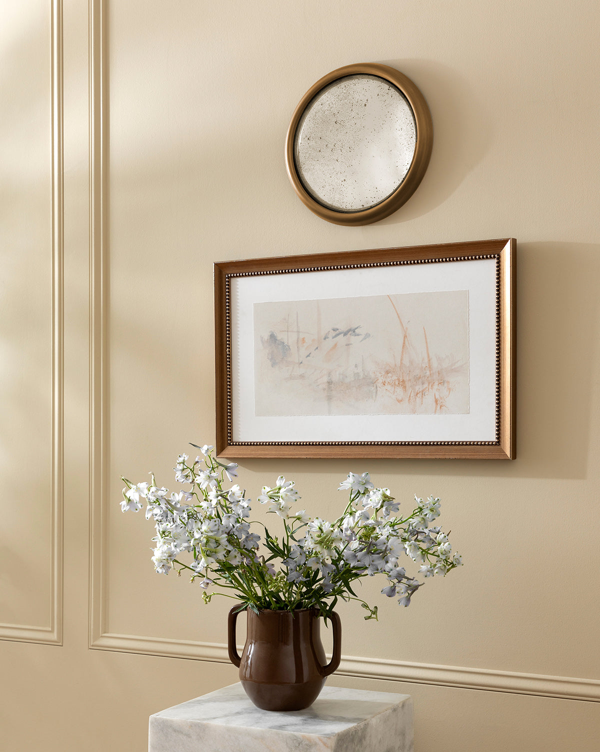 A brown vase with white flowers sits on a marble pedestal beneath the Juliette Convex Antiqued Mirror and a round decorative plate, all set against an antique-inspired beige paneled wall, by McGee & Co.