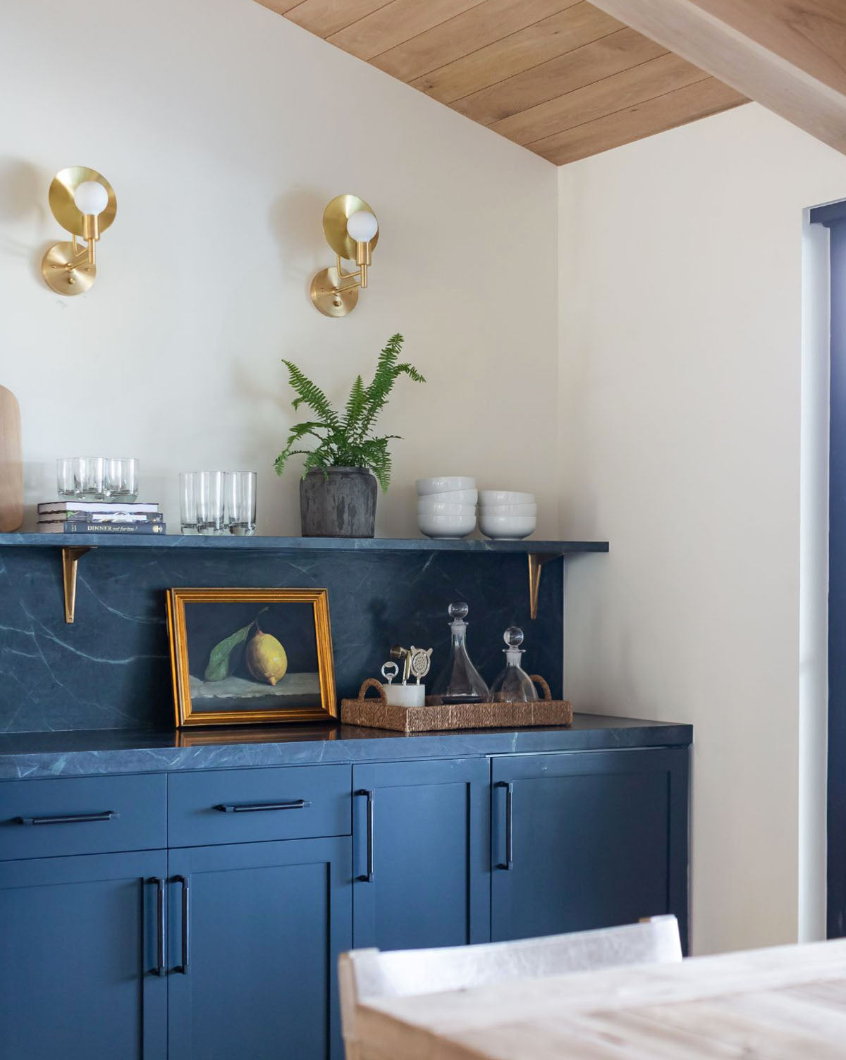A modern kitchen with navy cabinets, black marble countertop, gold sconces, a potted fern, white ceramics, glassware, and the Still Life with Lemon framed portrait on a floating shelf against a light wall.
