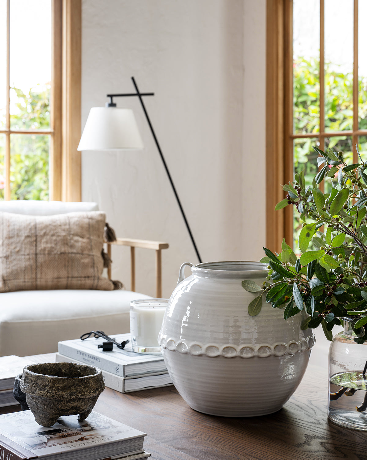 A bright living room with large windows features a cushioned chair with a pillow, a floor lamp, and a wooden table displaying the Rounded White Vase, a plant, books, a candle, and a small stone bowl.