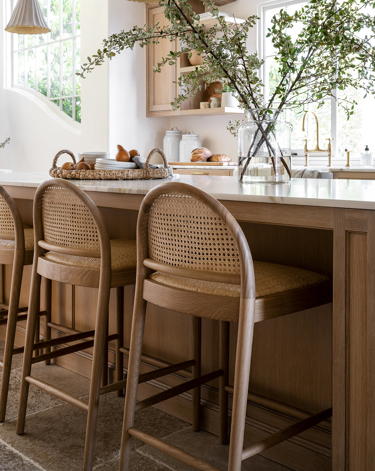 A bright kitchen features light wood cabinets, a marble island, and three Hadden Natural Counter Stools with oak frames. Sunlight streams through the window, illuminating a vase with green branches and a woven bread tray on the counter by McGee & Co.