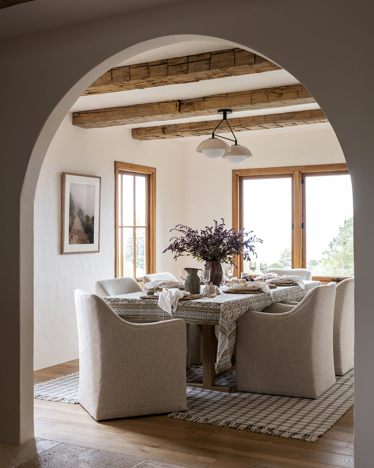 A cozy dining room features beige upholstered chairs, a wooden table with a patterned tablecloth and purple flowers, large windows, wood beams, gentle natural light—and “Foggy Path” giclee print enhancing the wall.