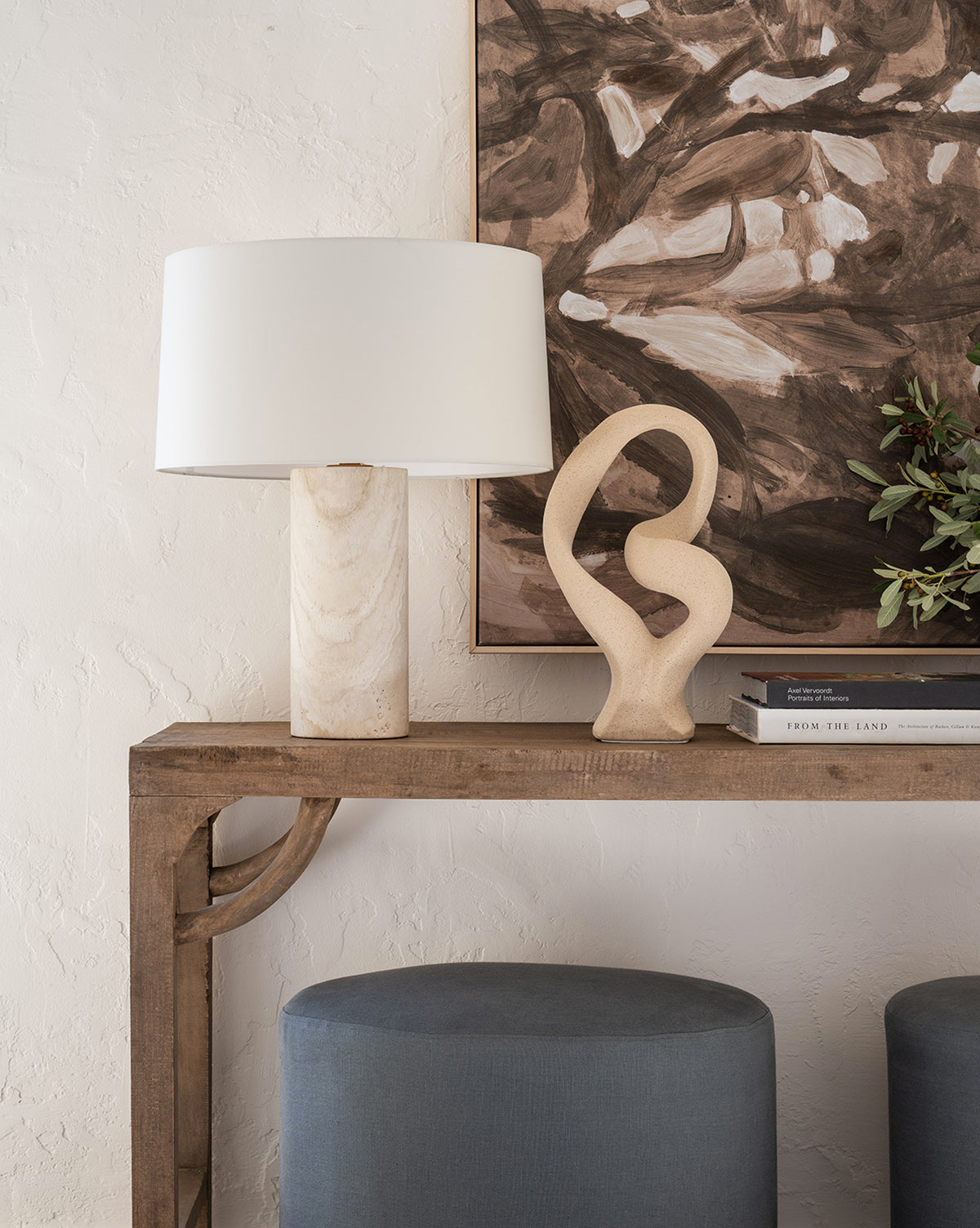 A wooden console table displays the Vespera Table Lamp with a marble base and brass accents, set next to an abstract beige sculpture, stacked books, greenery, two blue ottomans below, and a bold brown-and-white painting overhead by McGee & Co.