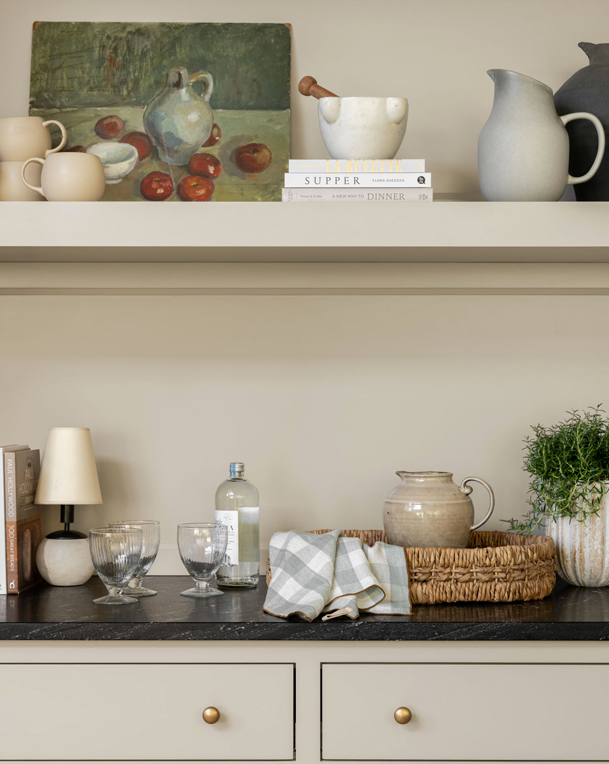 A neutral-toned kitchen counter holds glasses, a bottle, the Levi Stoneware Pitcher, a woven basket, a potted plant, checked cloths, and books. Pottery and a still-life painting are displayed on shelves above by McGee & Co.