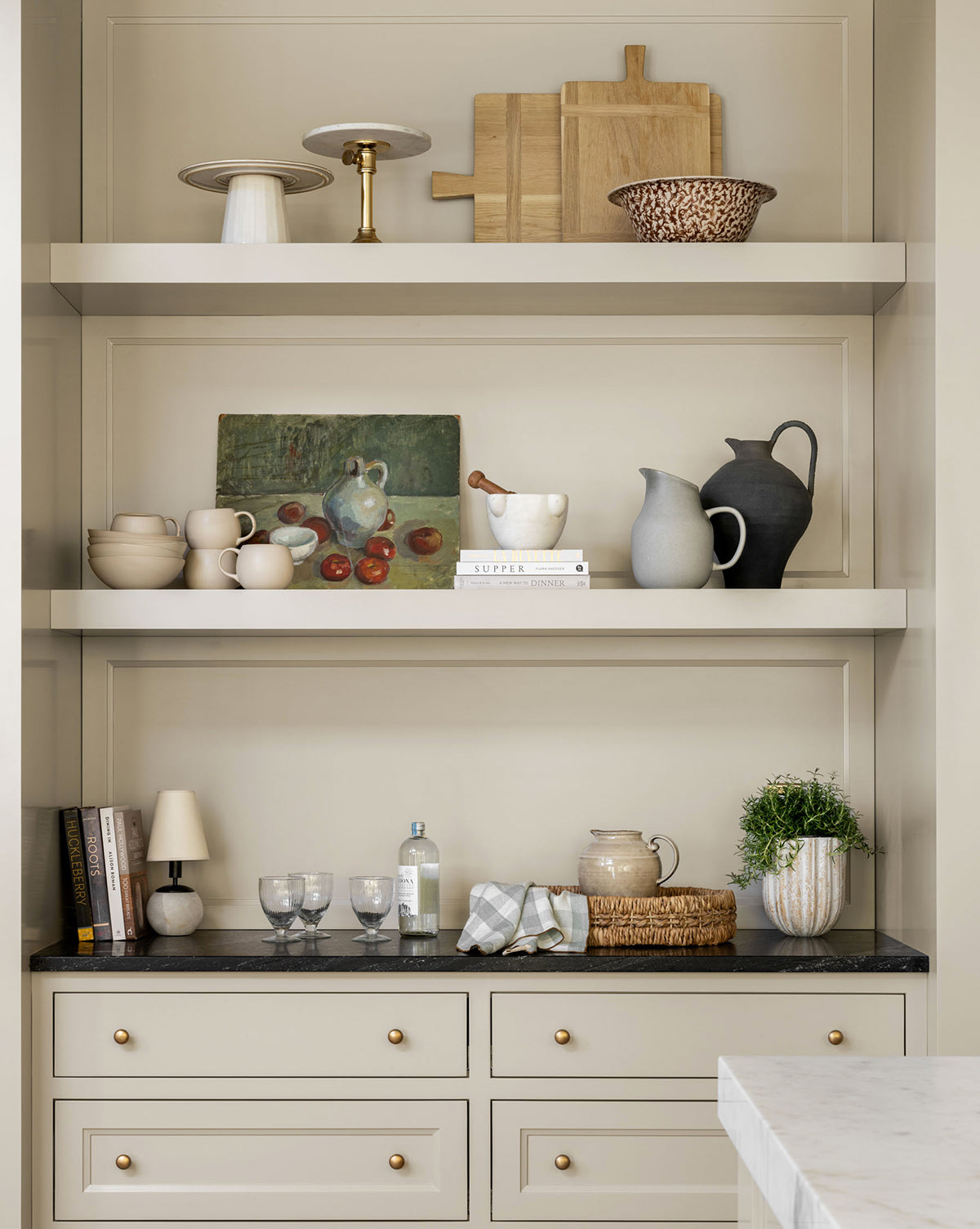 Open shelves with neutral dishware, glassware, a Rectangle Bread Board in a natural finish, oak and walnut cutting boards, a plant, lamp, books, bottle, and a small framed fruit painting—all atop cream cabinetry with gold knobs by McGee & Co.