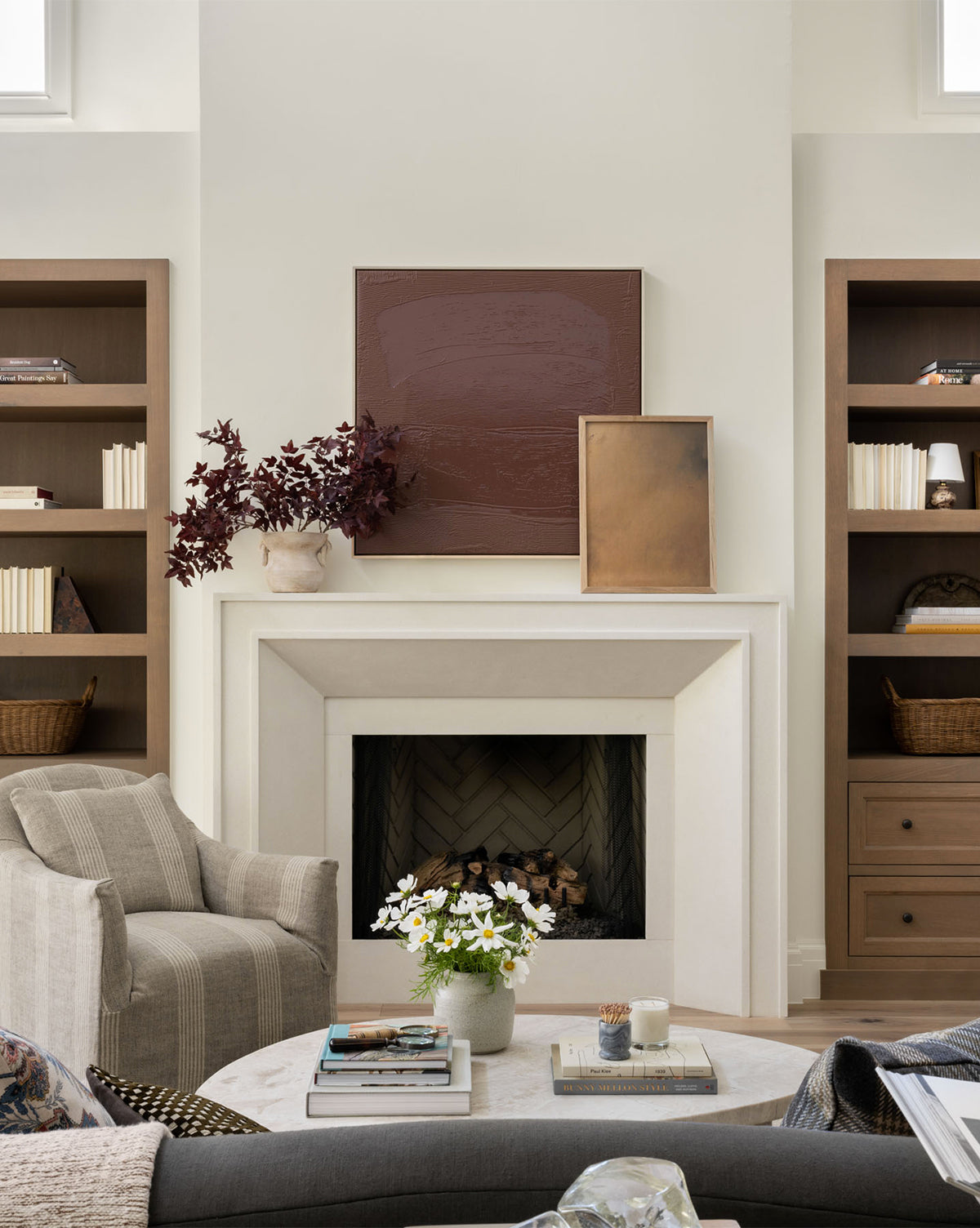 A cozy living room featuring a light-colored fireplace, elegant decor, two wooden bookshelves, a gray armchair, and a round coffee table with flowers and candles. The Golden Hour abstract wall art is displayed above the fireplace.