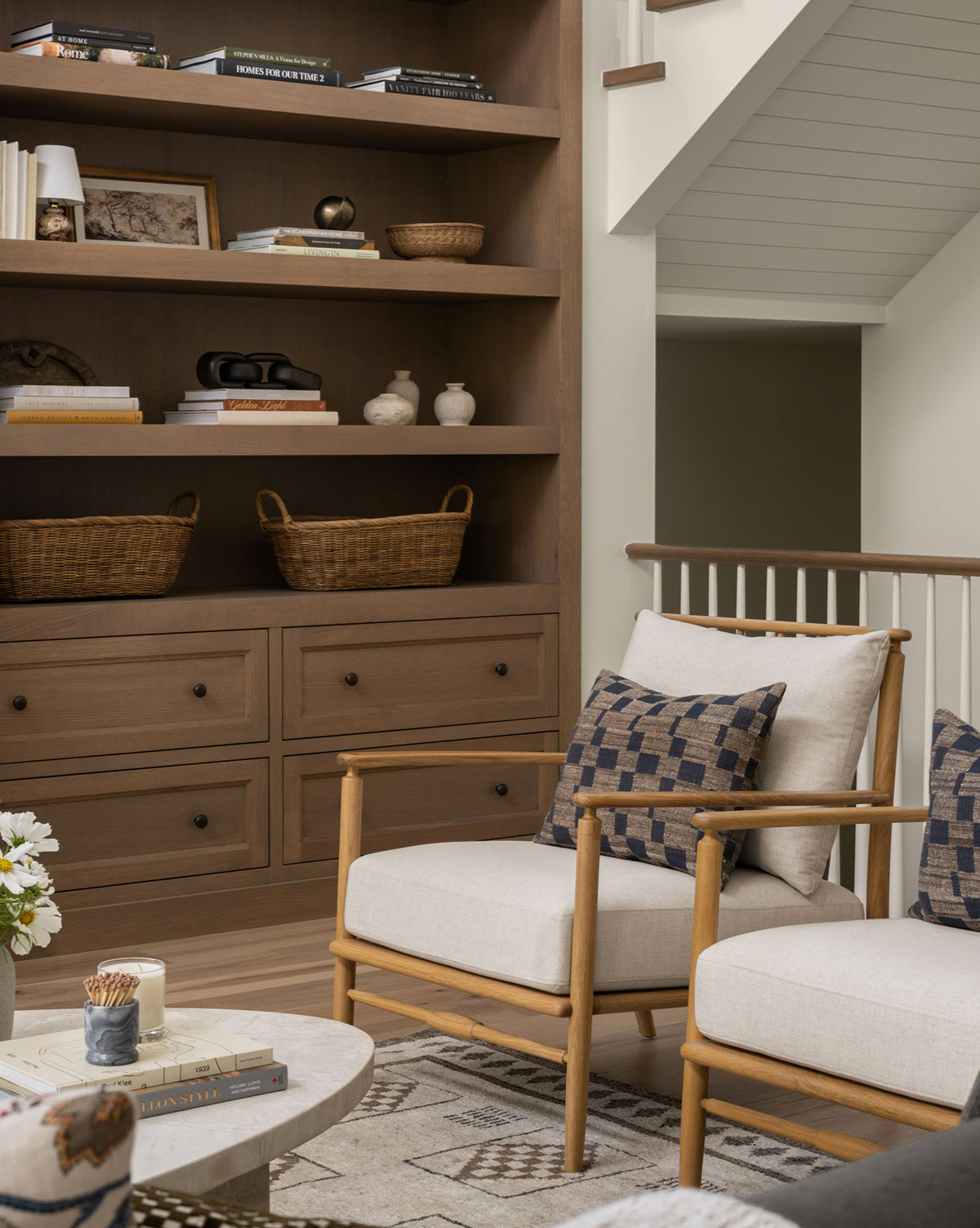 A cozy living room corner features solid oak built-in shelves, baskets, books, decorative objects, Fenwick Lounge Chairs with linen cushions, a round marble coffee table, and a soft neutral palette by McGee & Co.