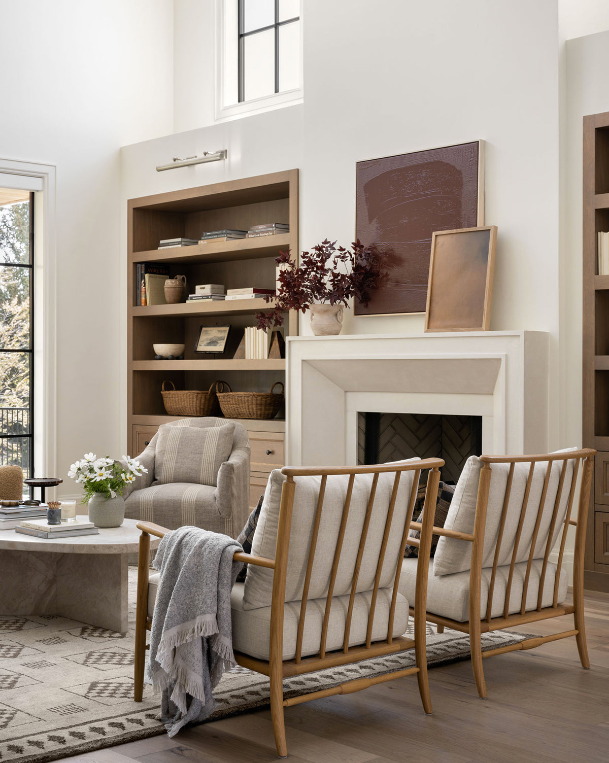 A modern living room featuring light wood furniture, Fenwick Lounge Chairs with linen cushions, a marble coffee table, built-in shelves with decor, a fireplace, and large windows that fill the space with natural light by McGee & Co.