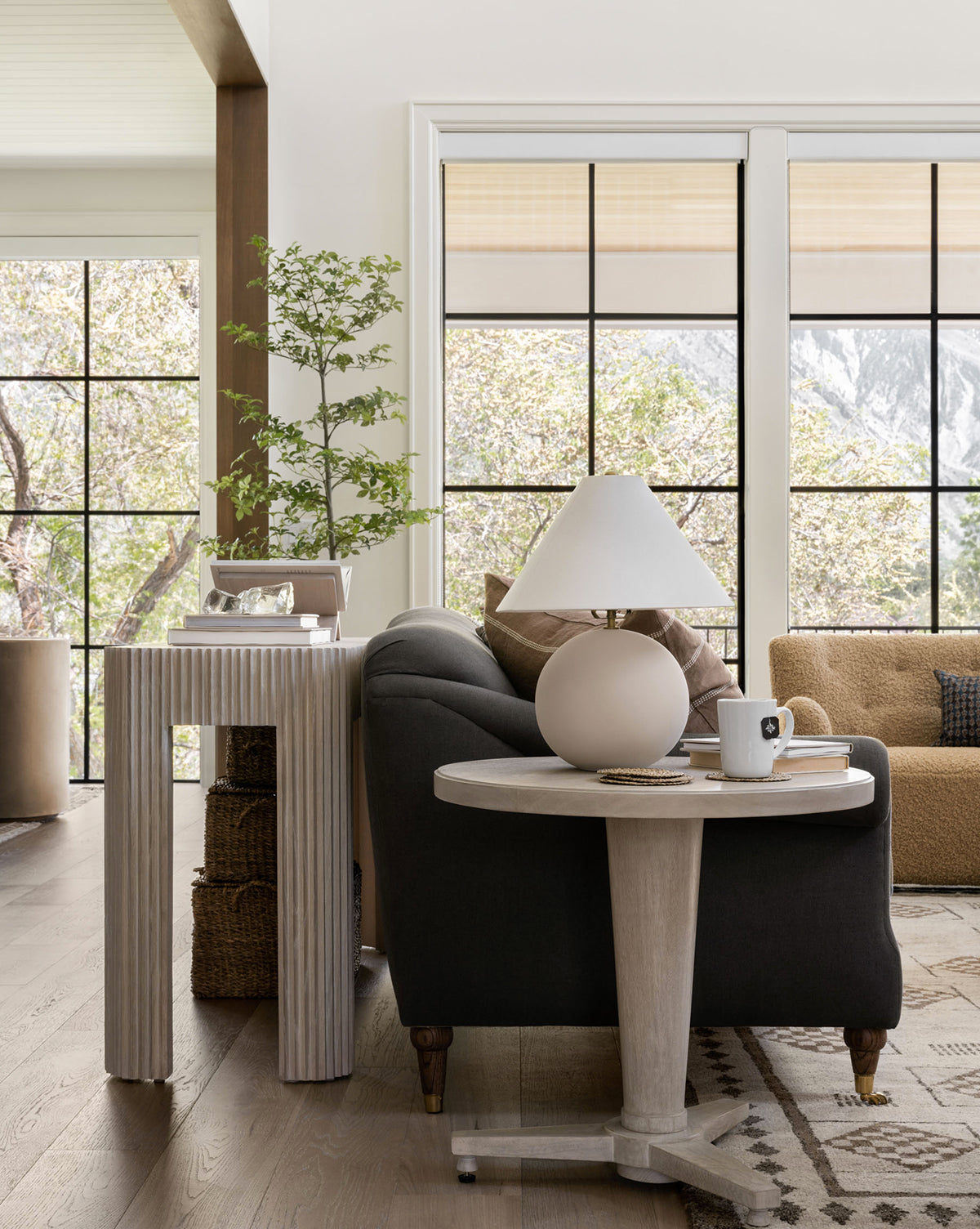 A cozy living room with a dark sofa, round light wood side table holding the Tess Ceramic Table Lamp and a coffee cup, large windows showing trees outside, plus a tan sofa and potted plant in the background by McGee & Co.