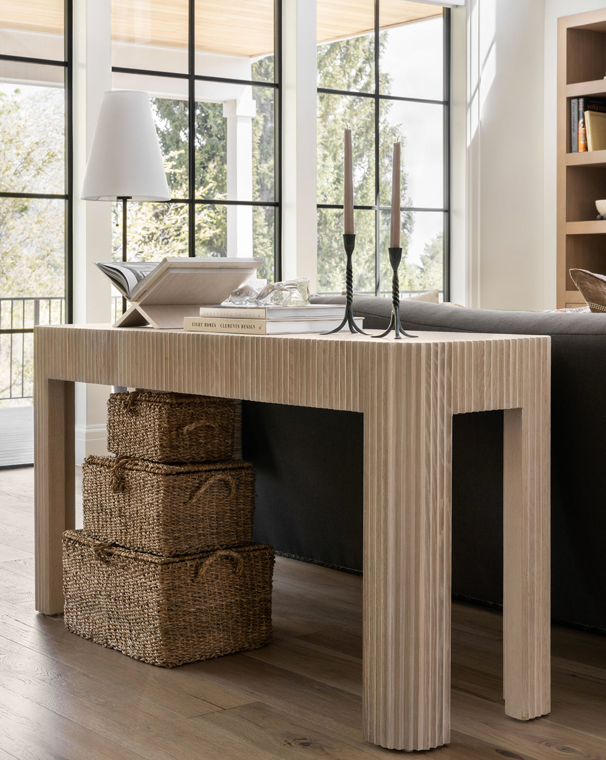 A light oak console table with fluted legs stands behind a dark sofa. On top are books, a Wooden Book Display, two black candlesticks, and a white lamp. Three woven baskets sit below as large windows fill the room with natural light.