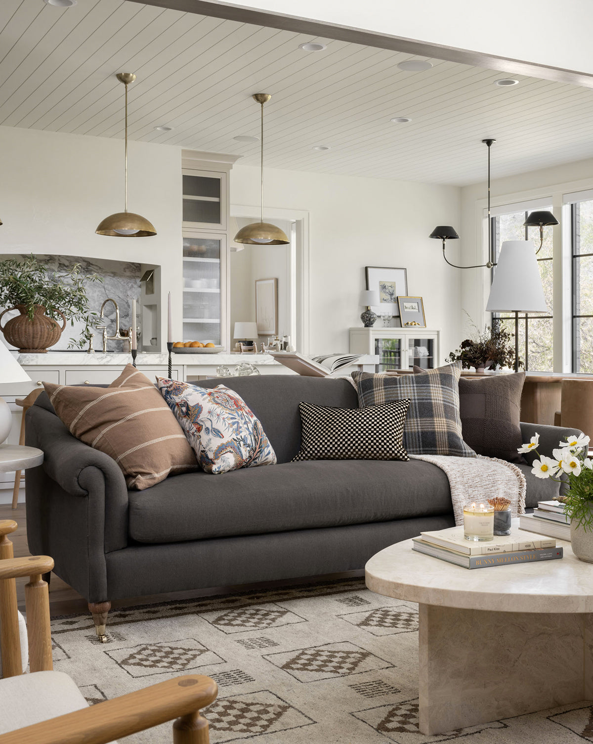 A cozy living room showcases the handcrafted Hildegard Sofa 90 in dark gray with patterned pillows, a round marble coffee table with flowers and candles, and an open kitchen and dining area, by McGee & Co. Neutral tones create a warm atmosphere.