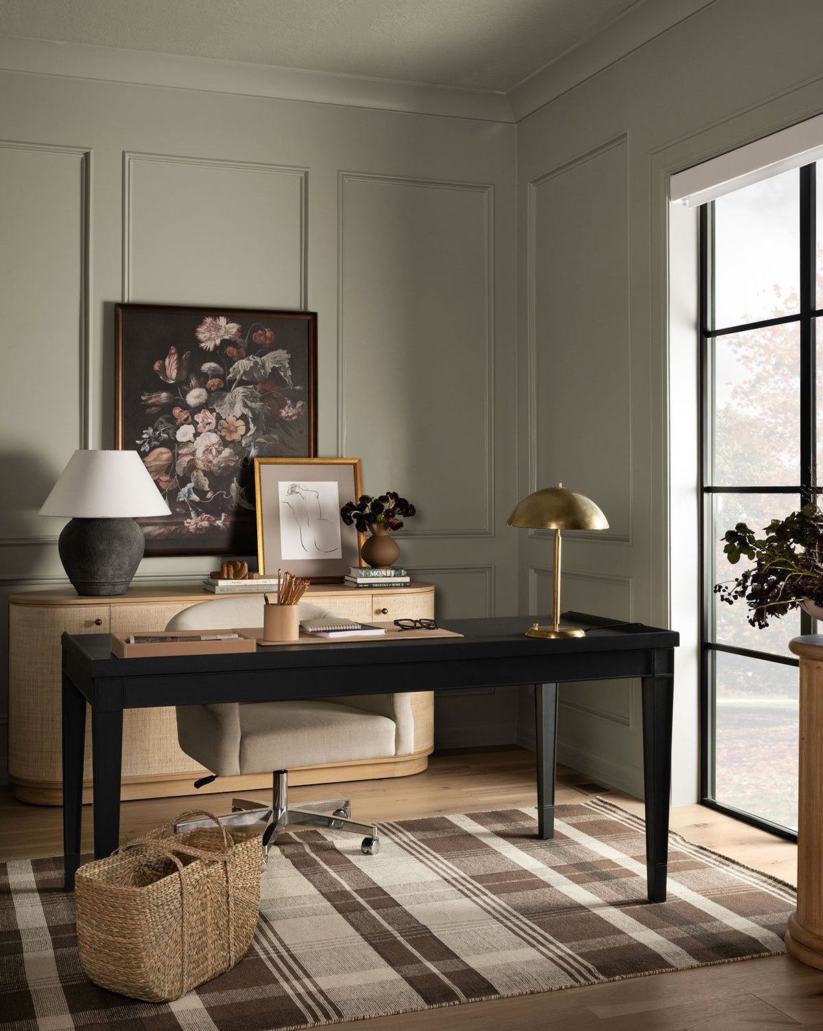 A modern home office with a black desk, beige chair, two lamps, framed floral art, woven basket, and large window. The paneled walls add texture, while the floor features the Orton Handwoven Wool Rug in a plaid pattern by McGee & Co.