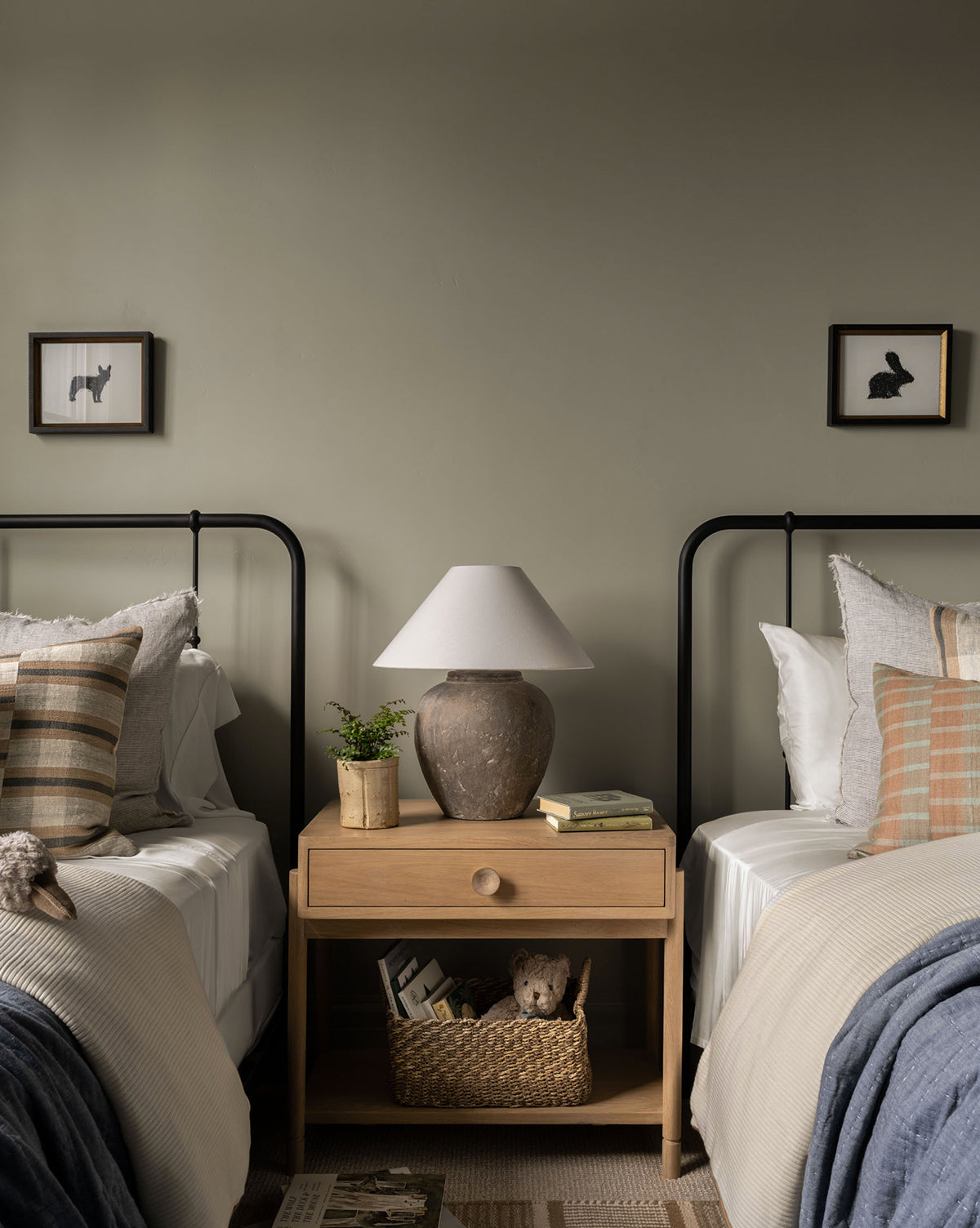 A cozy bedroom with two single beds, neutral bedding, decorative pillows, and a wooden nightstand in between. A lamp, books, and a plant sit atop the nightstand. Above each bed hangs Scribbled Rabbit linework wall art.