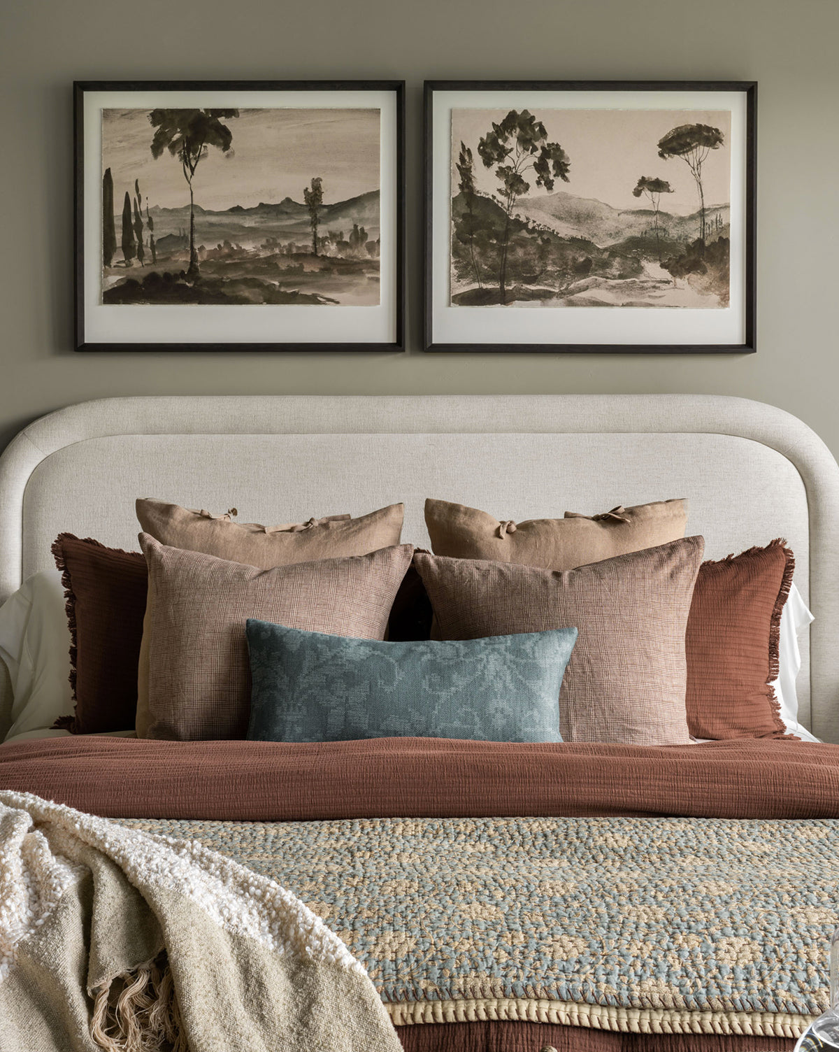 A cozy bed with brown and blue pillows sits against a light headboard. Above, two Washed Terrain II black-framed watercolor art prints hang, while a textured blanket is draped at the foot of the bed.