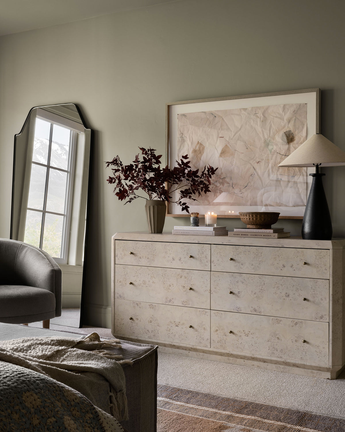 A light dresser with gold knobs displays a vase with dark red leaves, candles, a basket, and two lamps. The Giles Floor Mirror with a matte black frame leans against the wall by the window; abstract art is framed above the dresser, by McGee & Co.