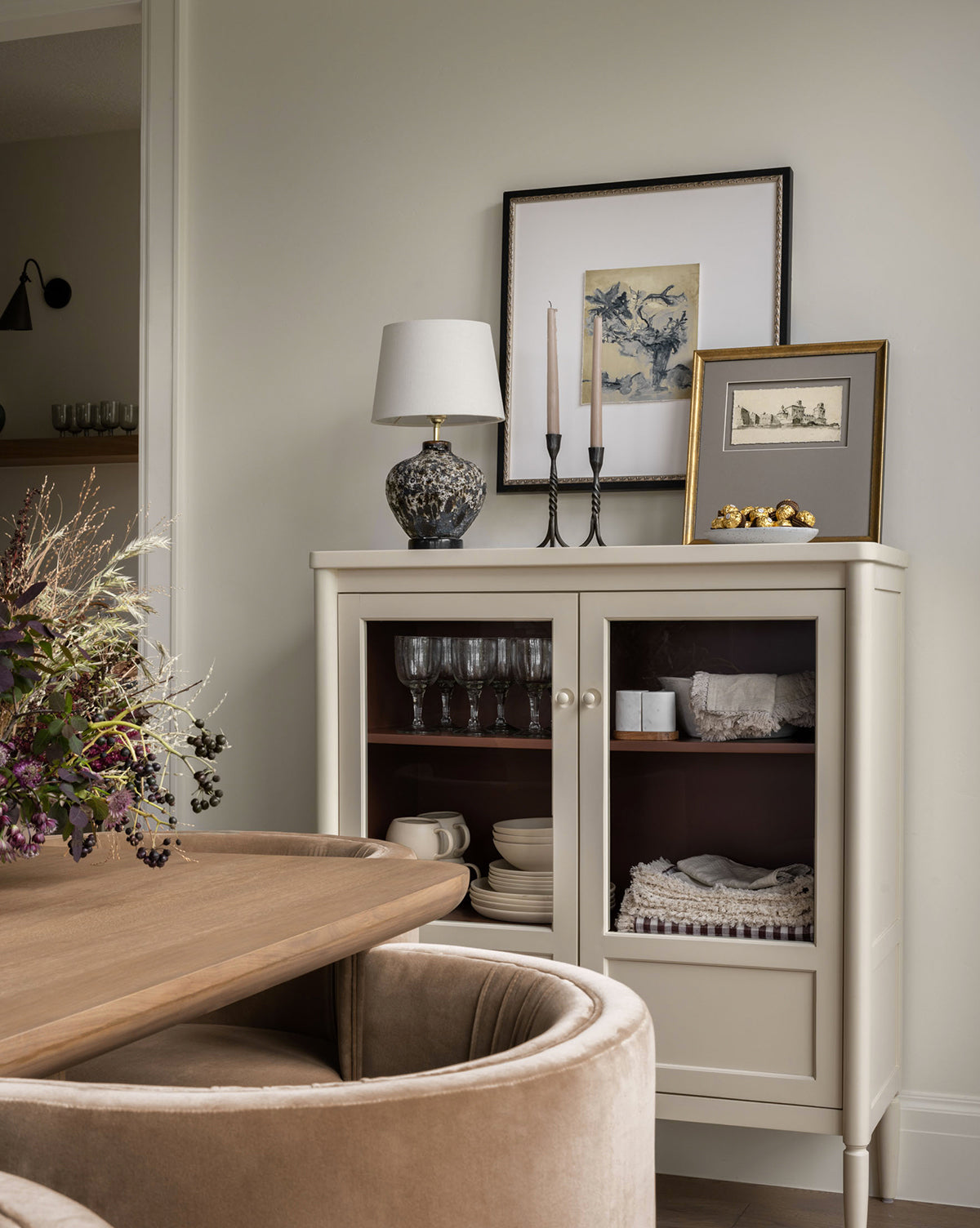 A cozy dining nook features a round wooden table, beige velvet chairs, and the Highgate Cabinet in bone white displaying dishes. Atop the cabinet sit framed art, a lamp, candles, and a bowl with gold ornaments by McGee & Co.