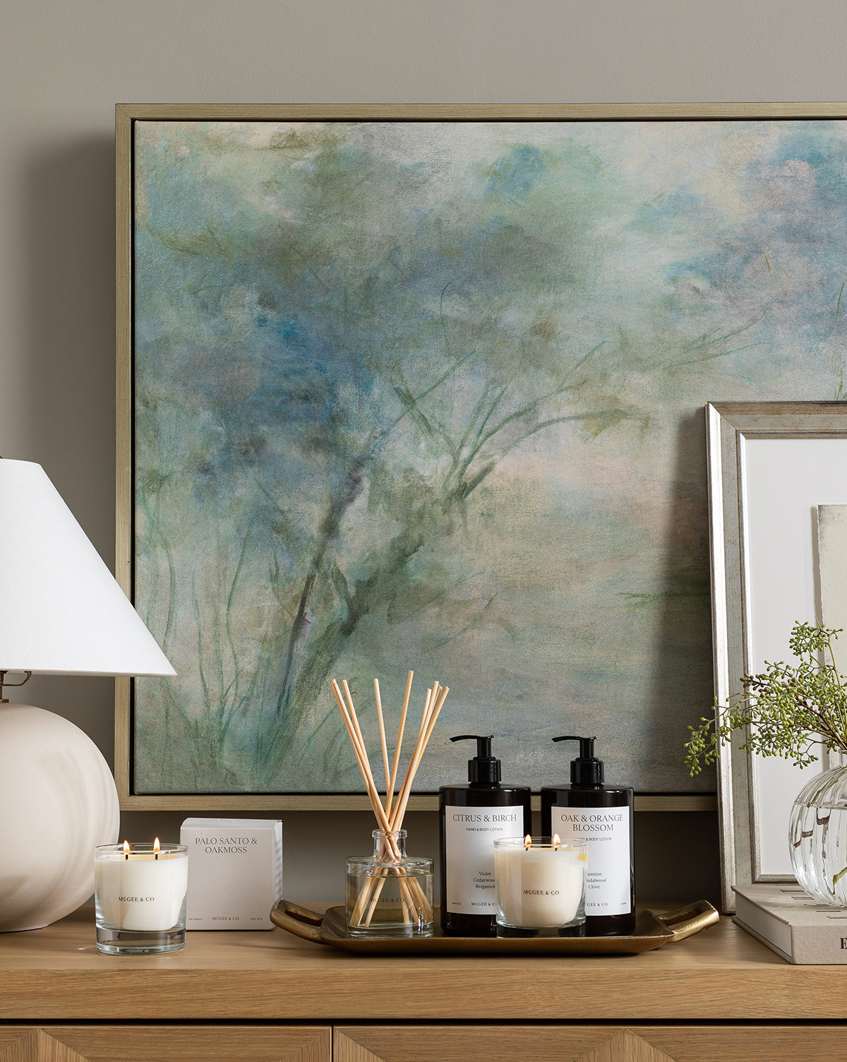 A serene bathroom countertop features Fading abstract blue-green wall art, a round white lamp, two lit candles, a glass reed diffuser, and bottles of hand wash and lotion arranged on a gold tray.