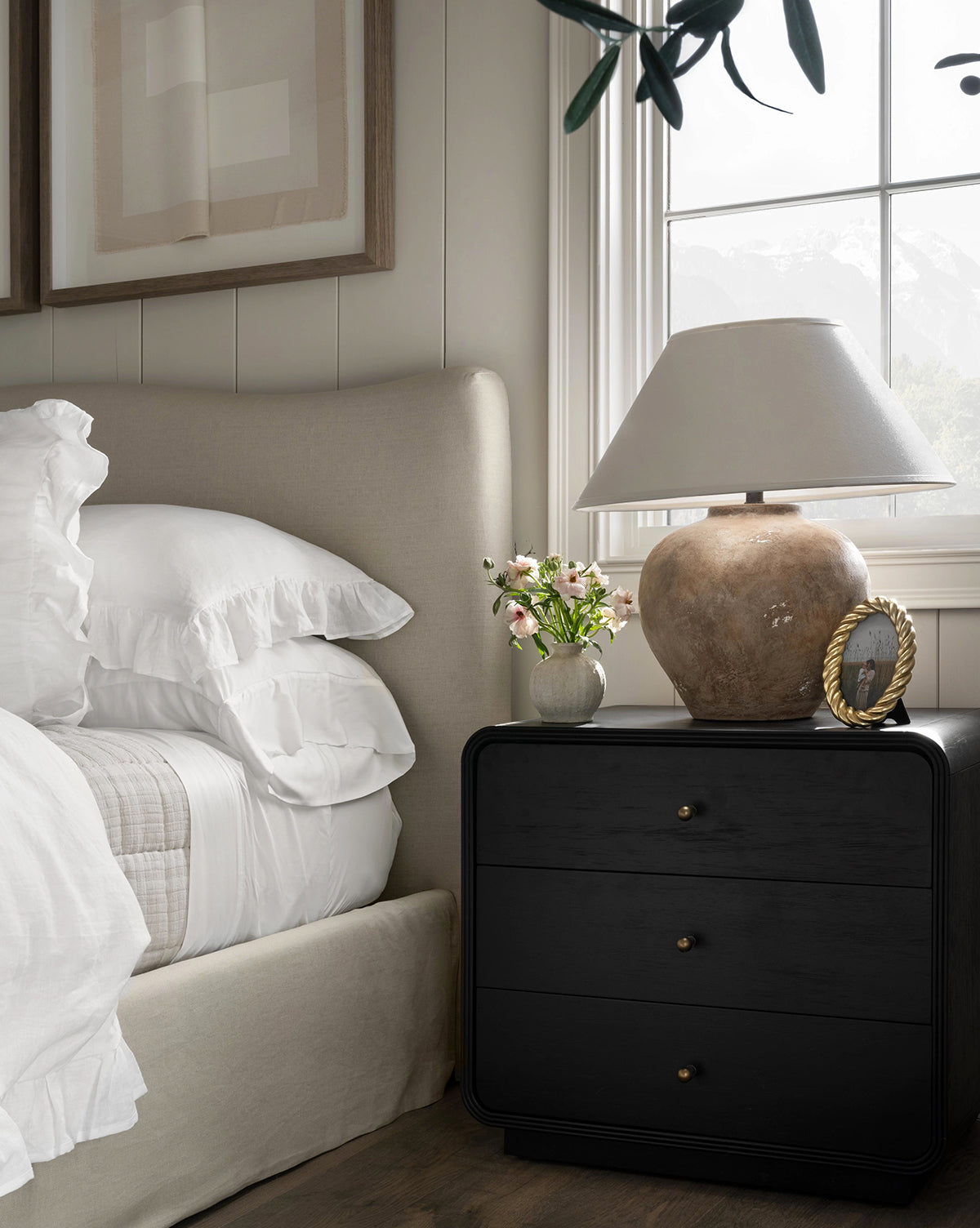 A cozy bedroom with a beige upholstered bed, white French Linen bedding, and a black nightstand topped with a Pura x Studio McGee French Linen Home Fragrance Refill, flowers, and a photo frame by a window framing mountain views.