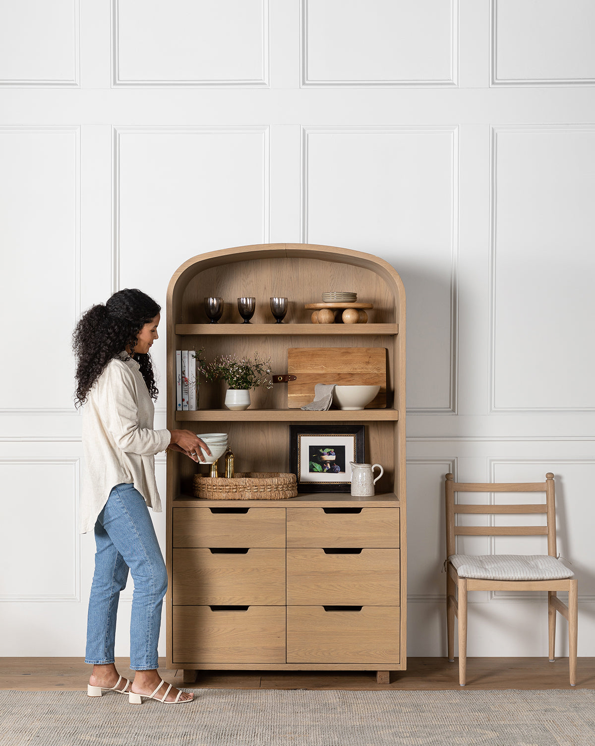 A woman arranges items in the Burnham Cabinet, which features shelves and drawers for storing books, glasses, a framed photo, and ceramics. Nearby, a cushioned wooden chair sits by a white paneled wall—ideal for dining room storage by McGee & Co.
