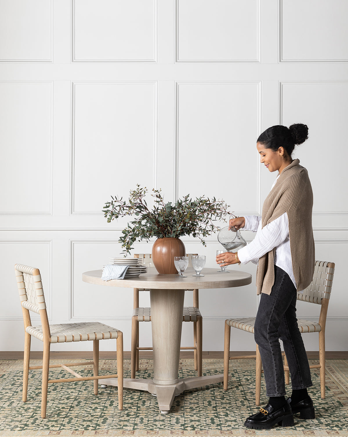 A woman stands by the Orla Dining Table with a white finish, pouring water into a glass. Two chairs, books, and a brown vase with green foliage sit atop the table in an indoor setting with a white paneled wall and patterned rug by McGee & Co.