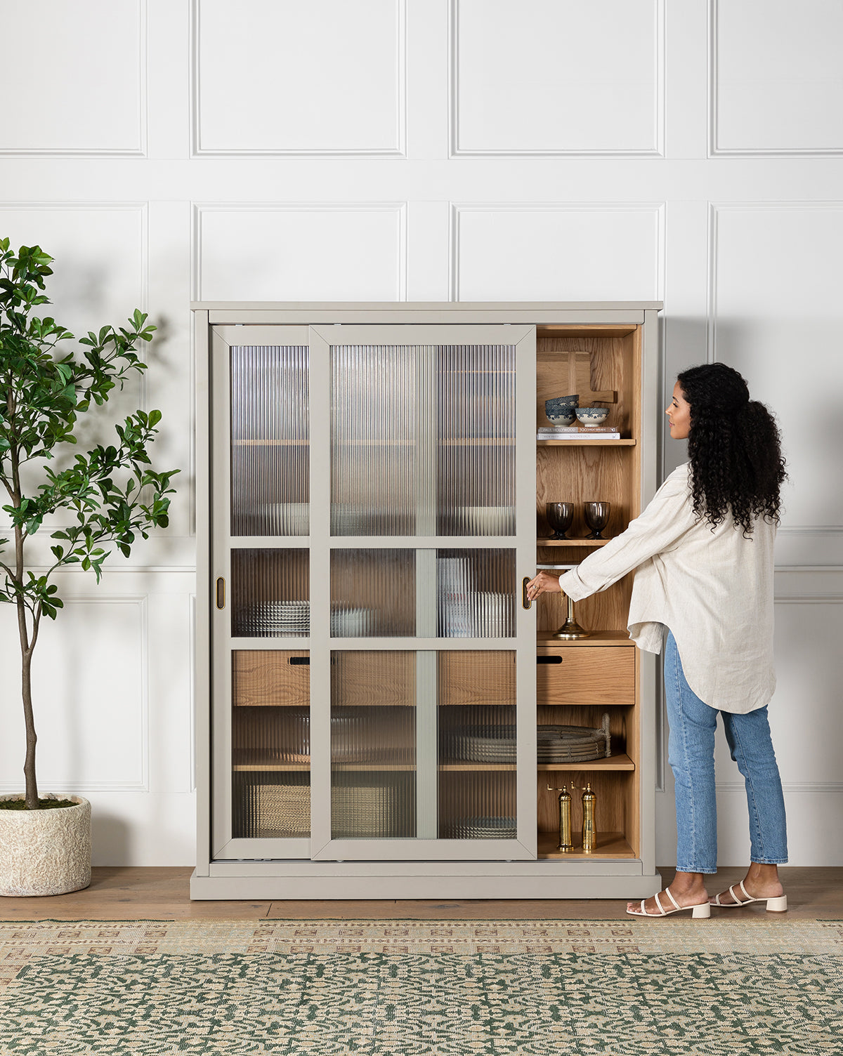 A woman opens the reeded glass doors of the Phillips Cabinet, a painted oak piece with shelves and drawers, by McGee & Co.