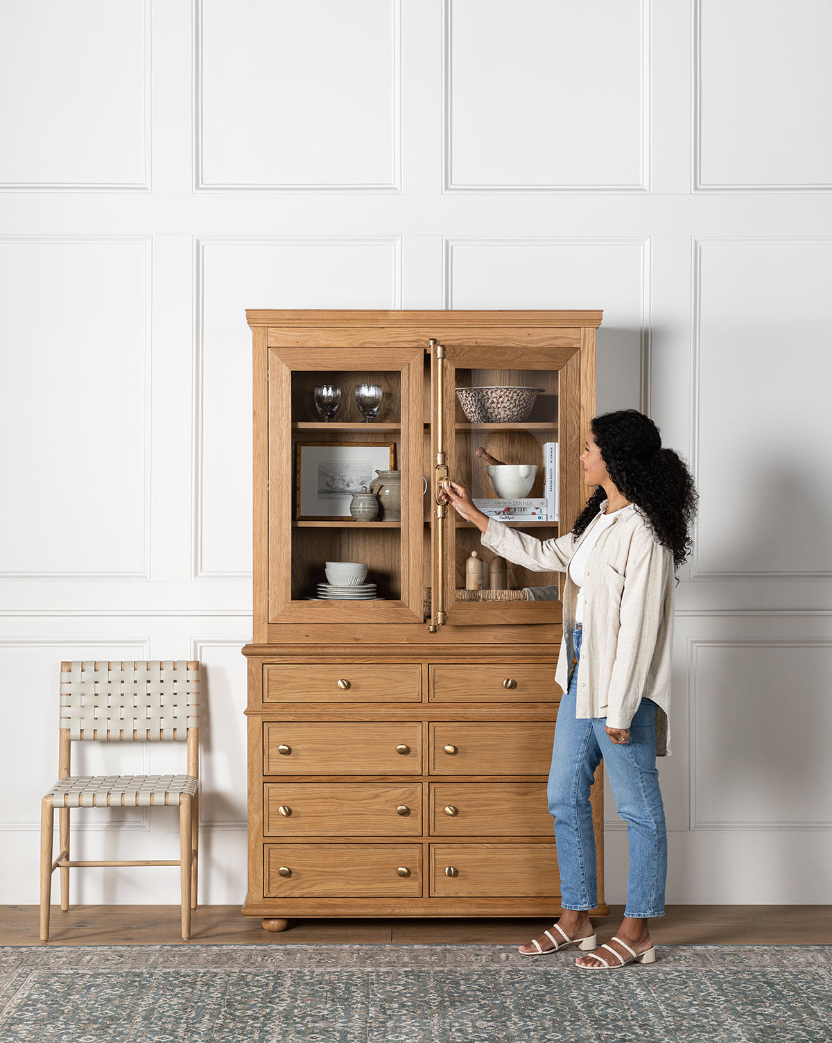 A woman opens the glass door of a Heath Oak Cabinet, revealing dishes and décor inside. Nearby, a woven chair sits against a white-paneled wall, accentuating the room’s traditional design by McGee & Co.