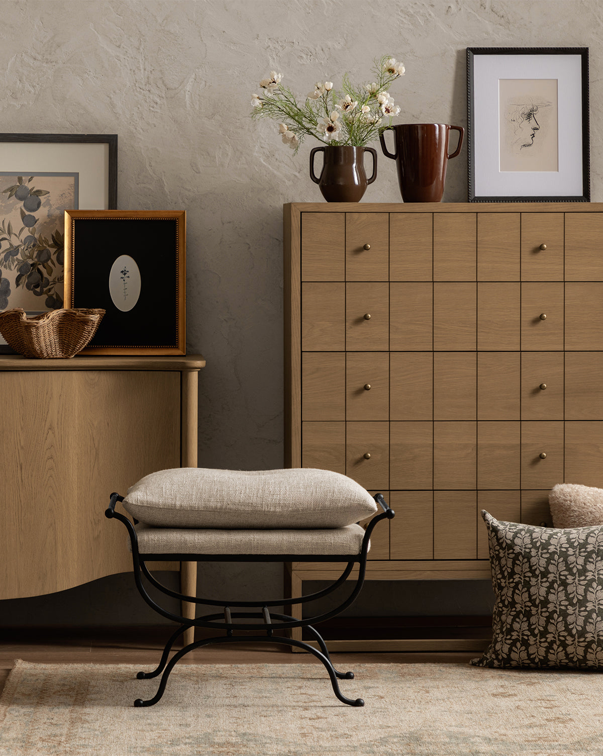 A cozy room with wooden furniture, a cabinet, and sideboard decorated with the Single Blooms 3 vase, flowers, a wicker bowl, and vintage wall art. A cushioned stool with a black frame sits on a patterned rug near a textured pillow, by McGee & Co.