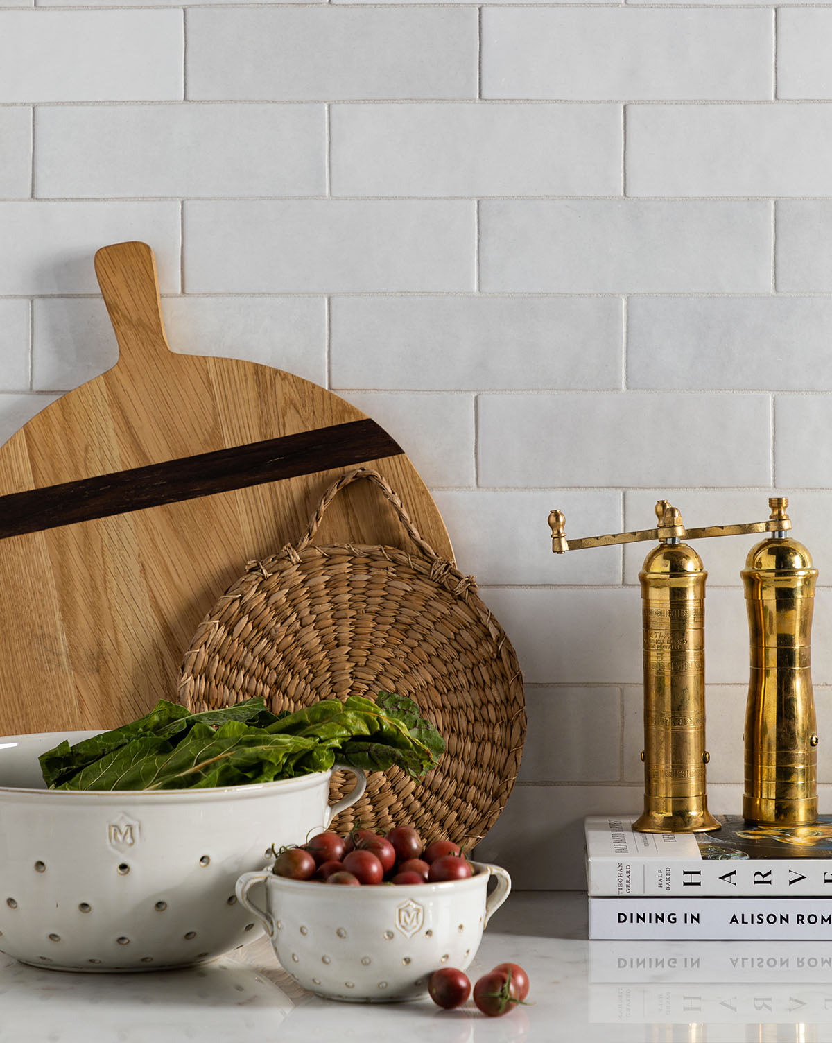 A kitchen counter with two Glazed Colanders of greens and cherry tomatoes, stylish serveware, a wooden cutting board, a woven trivet, brass pepper mills, and cookbooks against white subway tiles for chic decor, by McGee & Co.